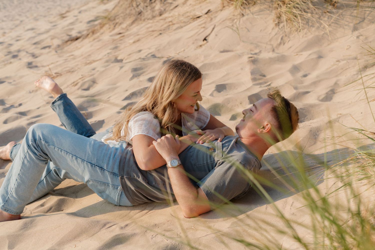 Couple lying together on sandy beach near dune grass during golden hour photoshoot.