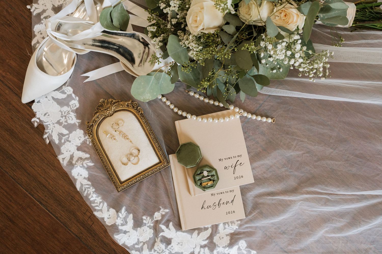 Wedding flat lay with white bridal heels, pearl necklace, rings, vow books, and a lush floral bouquet on lace fabric.