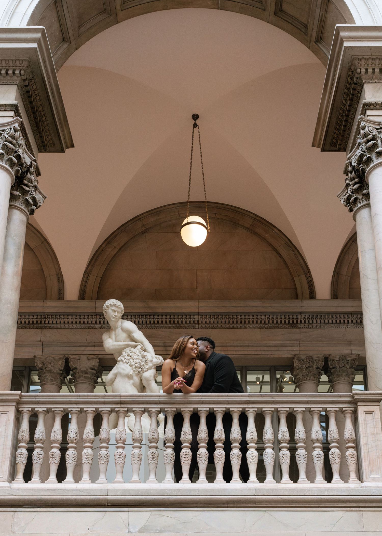 A couple shares an intimate moment on an ornate balcony with classical architecture and arched ceilings.