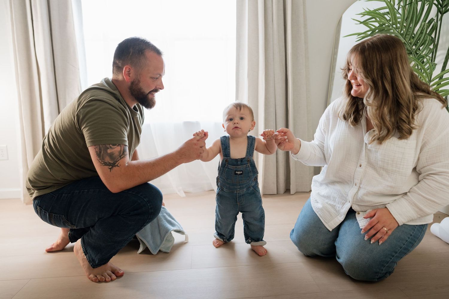 Parents helping their baby take first steps on hardwood floor in bright living room.