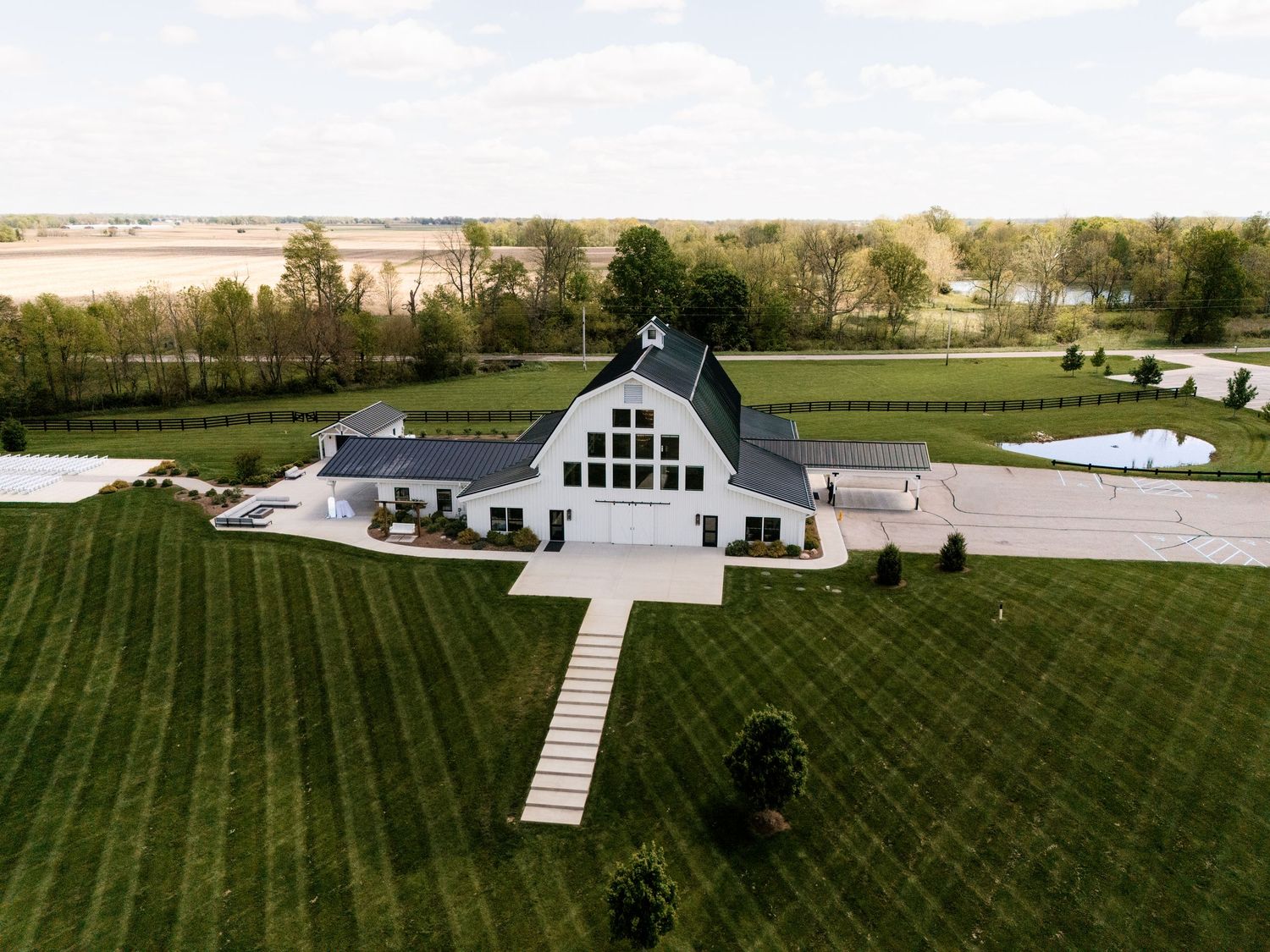 Aerial view of a elegant white barn wedding venue surrounded by lush green fields and manicured lawns on a sunny day.