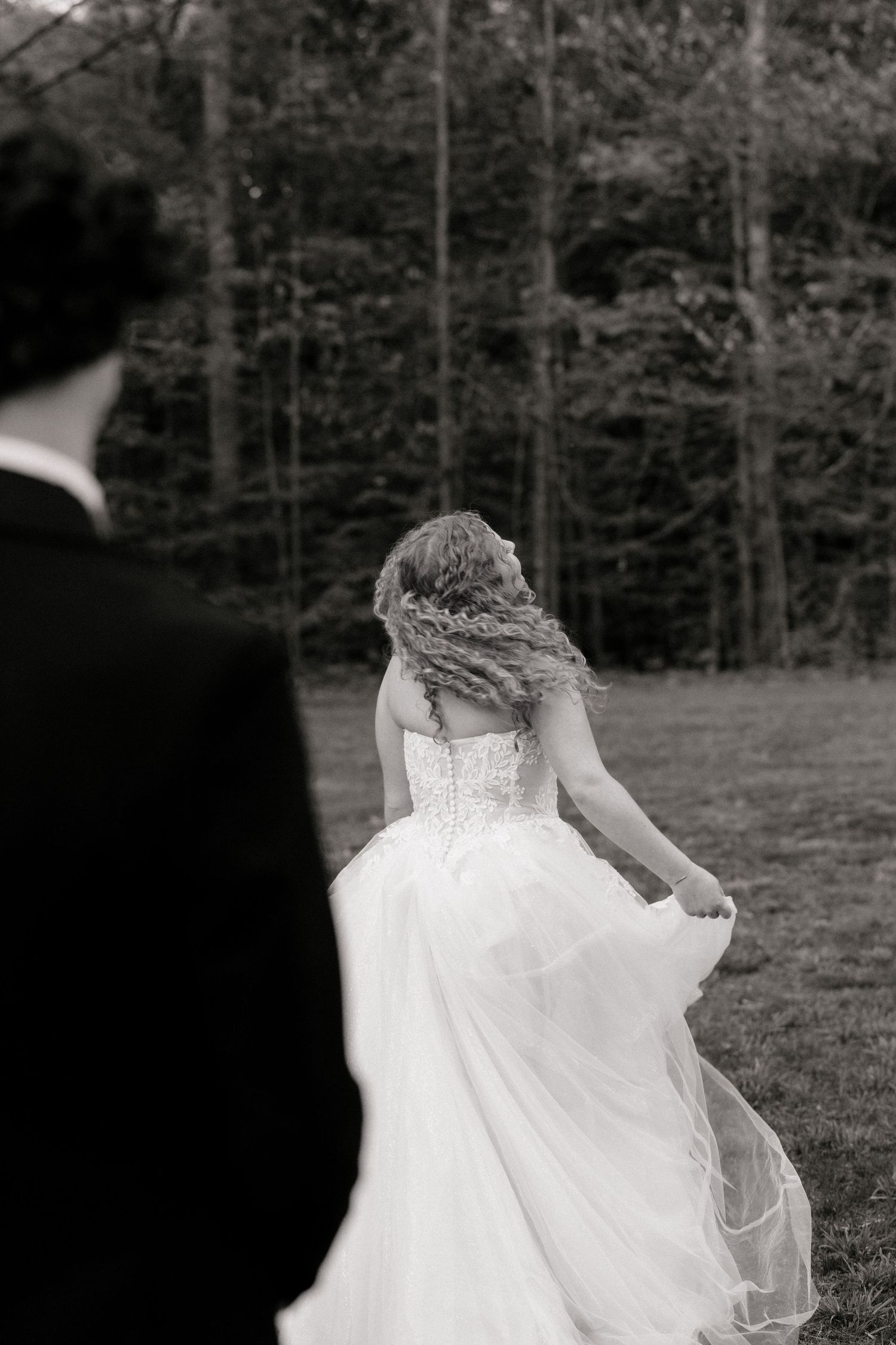 Black and white photo of a bride in a flowing gown twirling outdoors, with a groom in a suit watching her.