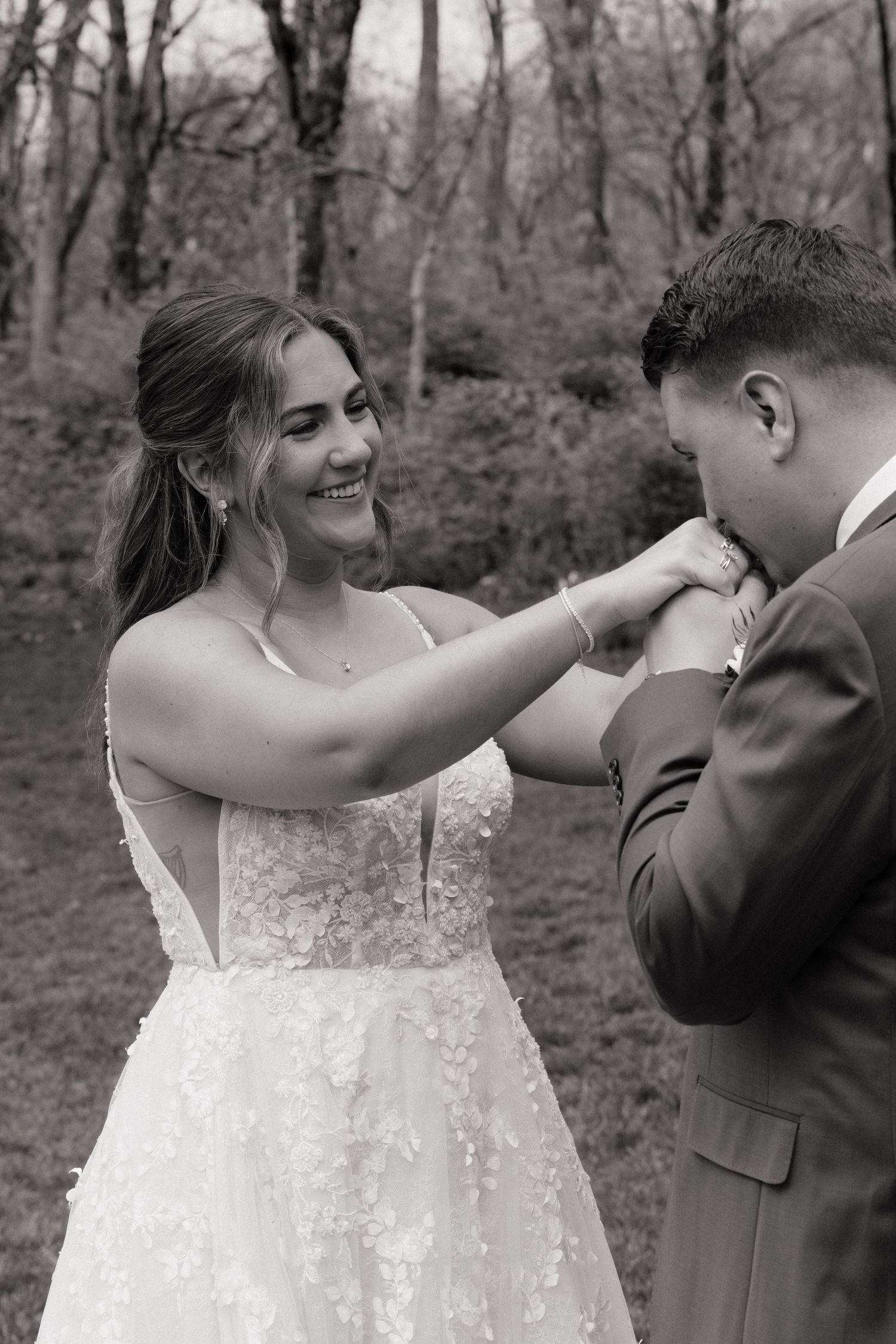 Black and white photo of a smiling bride in lace gown as groom kisses her hand outdoors.