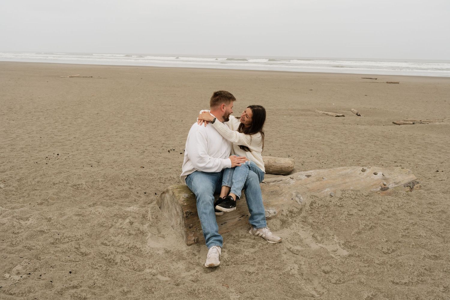 Couple sitting on driftwood log on sandy beach, embracing romantically on an overcast day.
