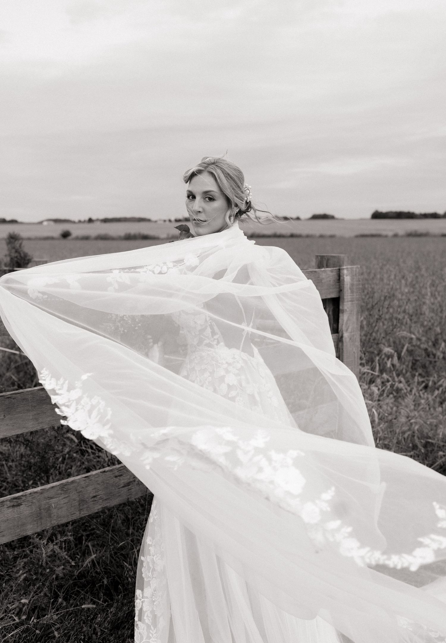 Black and white photo of a bride in a lace wedding dress with a flowing veil, posing by a wooden fence in an open field.