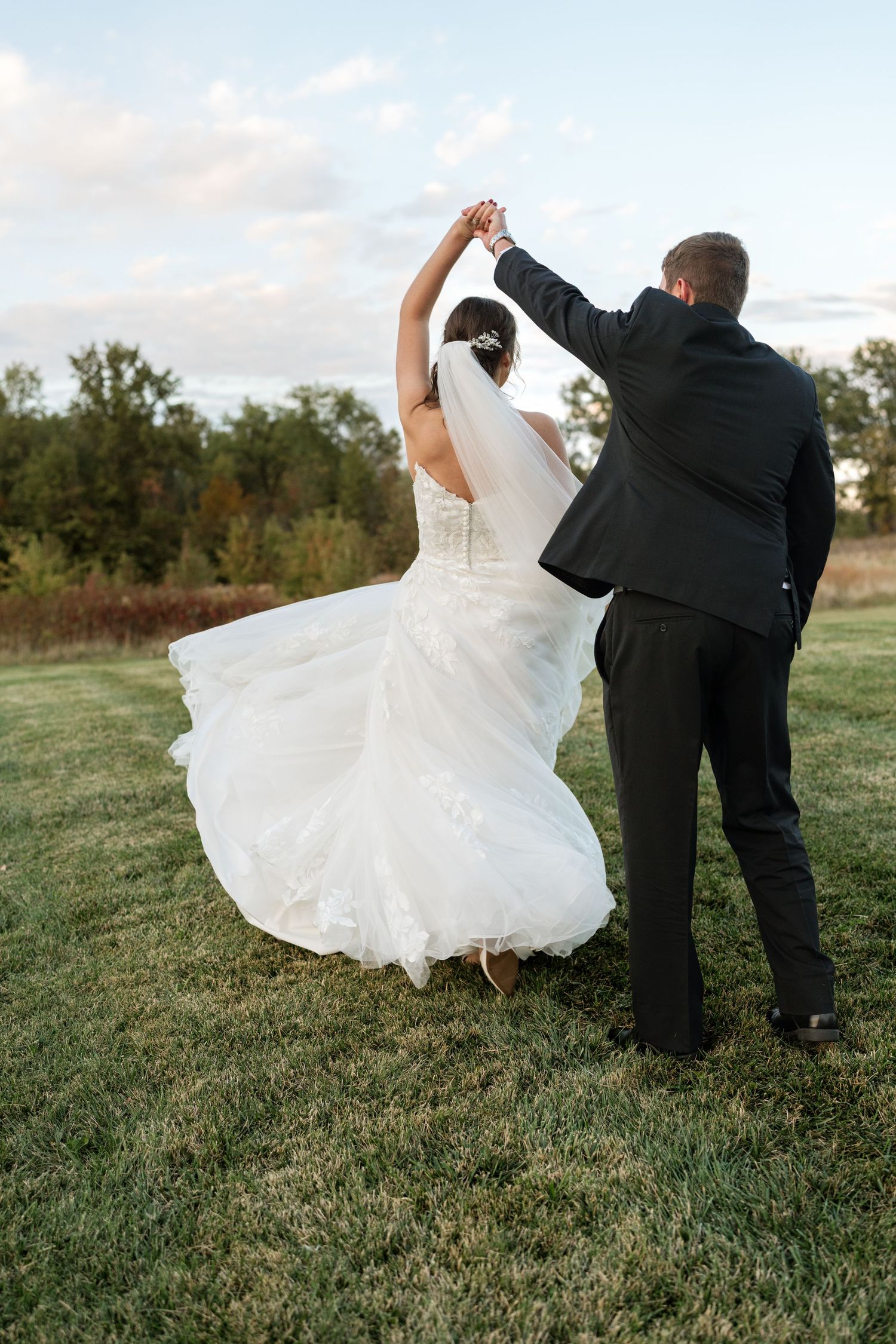 Wedding couple dancing and twirling on grassy hillside at sunset.