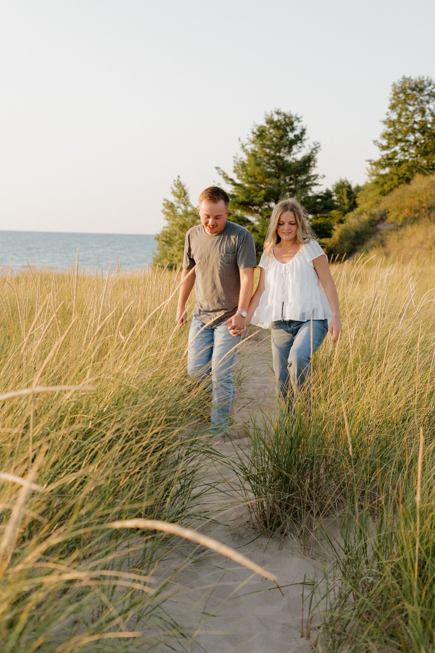Couple strolling through beach grass along sandy coastal trail with lake view.
