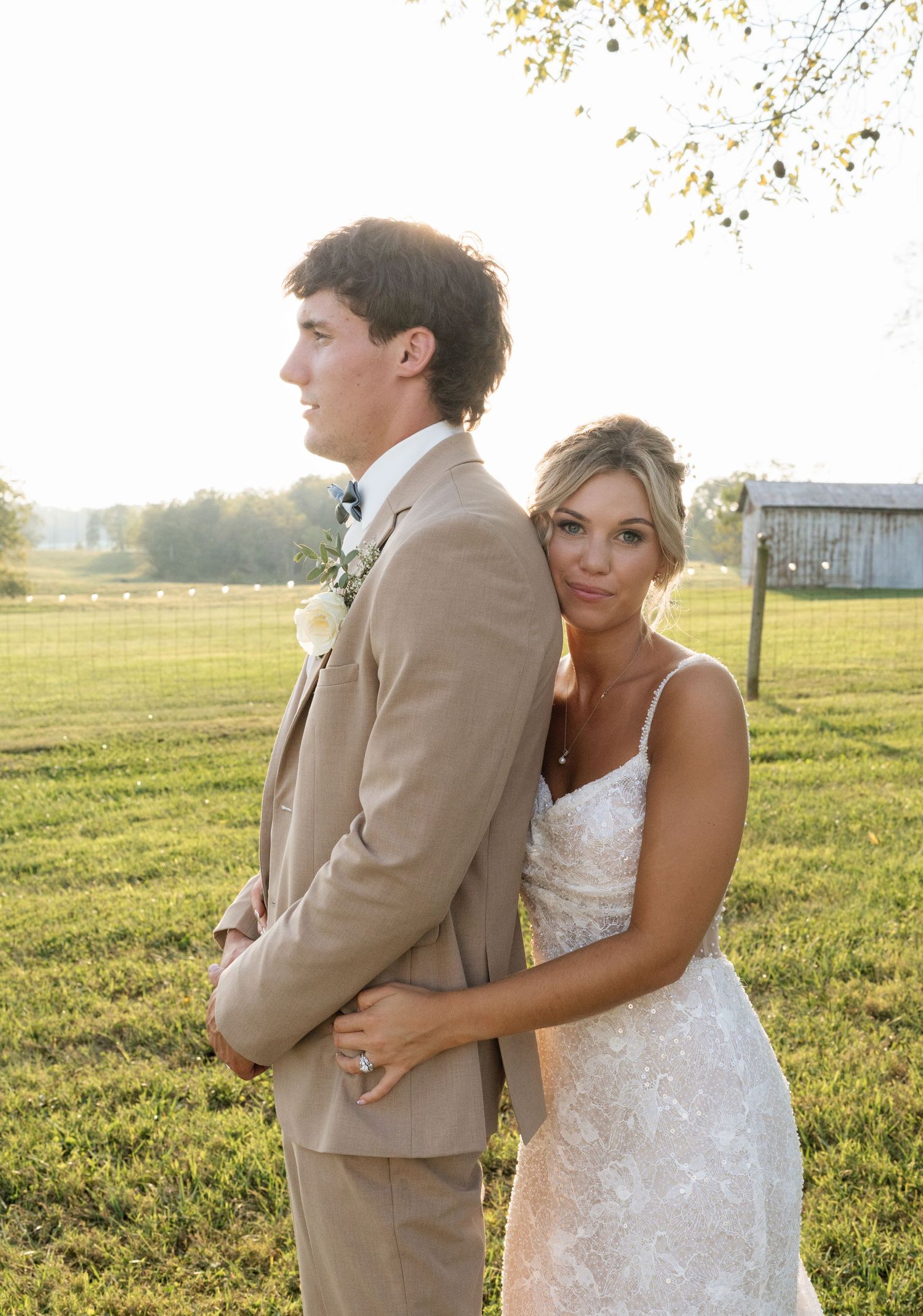 A bride in lace gown embraces groom in tan suit outdoors at golden hour on a rustic farm wedding venue.