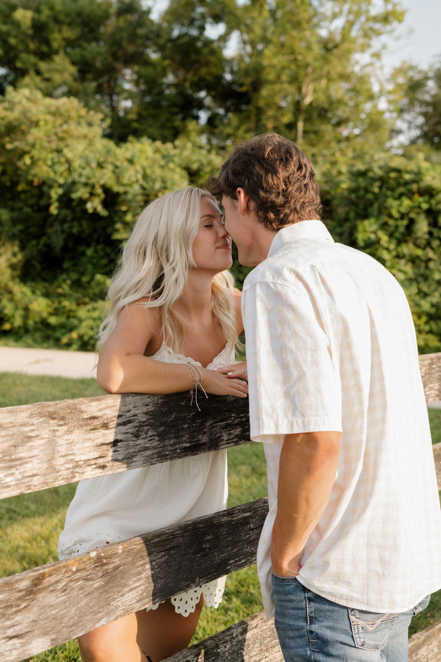 A couple shares an intimate moment by a rustic wooden fence in a sunlit outdoor setting.