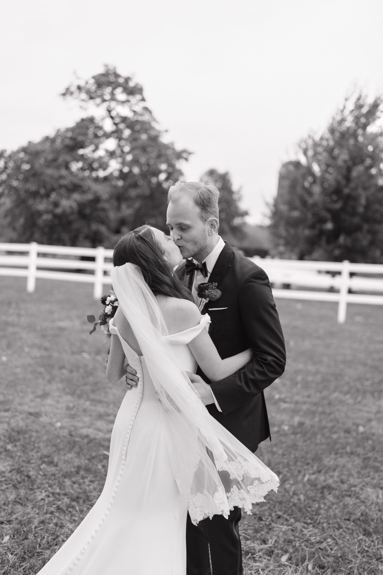 Romantic black and white wedding photo of couple dancing outdoors by white fence.