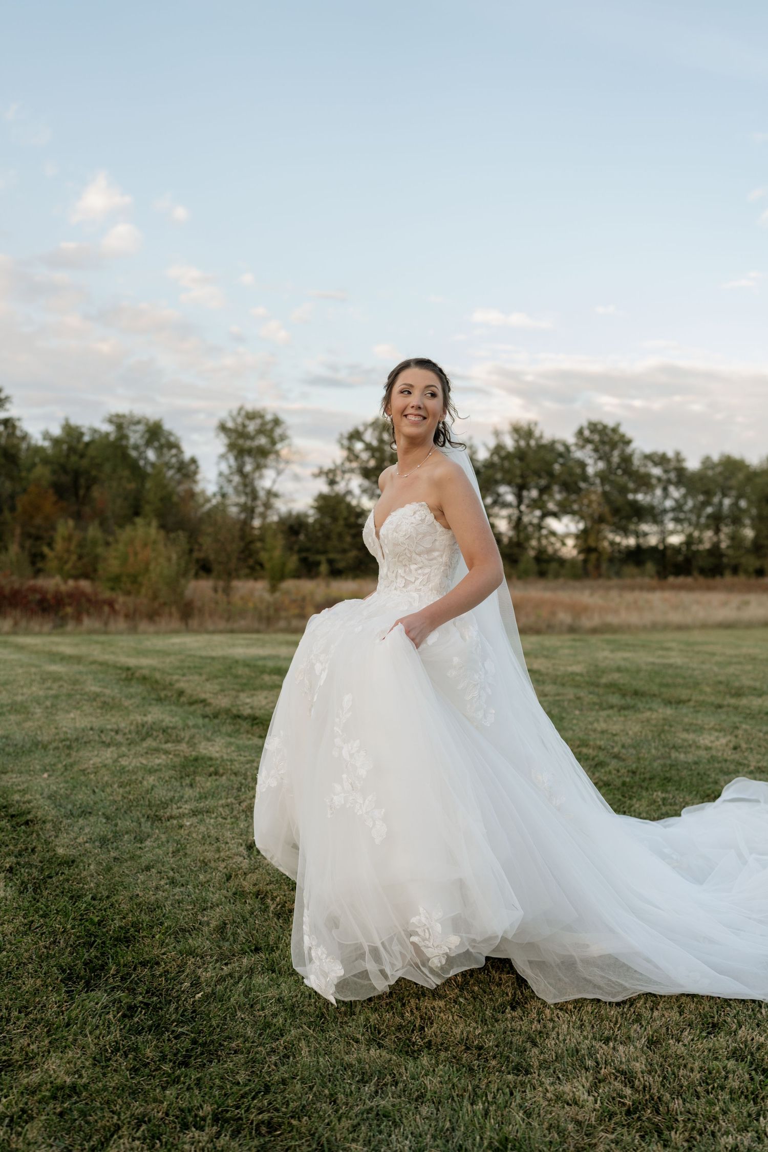 Bride in flowing white gown poses on grassy field during golden hour.