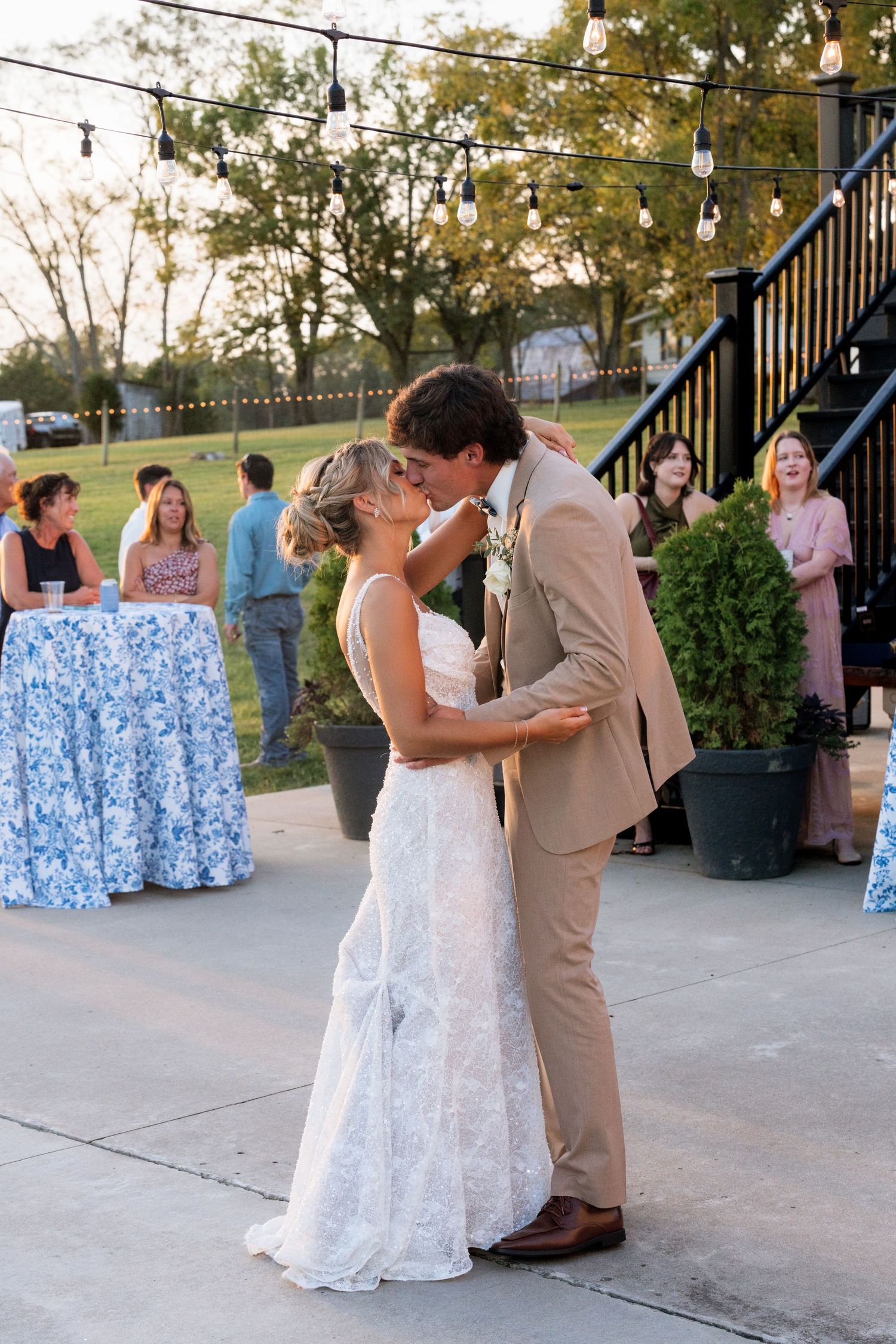 Bride and groom share first dance kiss outdoors at wedding reception with string lights and guests watching.