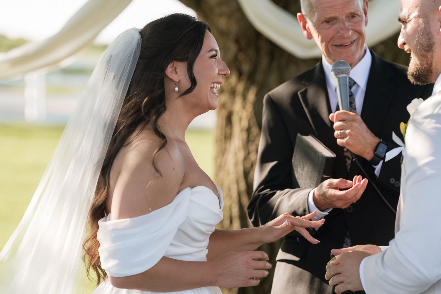 Bride in white off-shoulder gown smiling during outdoor wedding ceremony while officiant speaks into microphone.