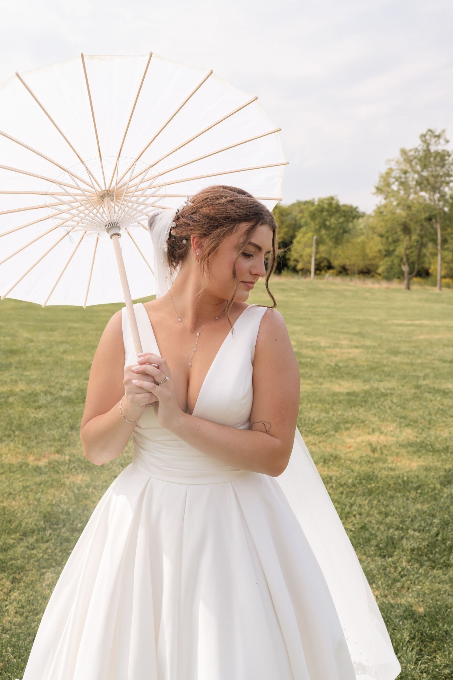 Bride in white gown holding a white parasol umbrella outdoors on green grass with trees in background.