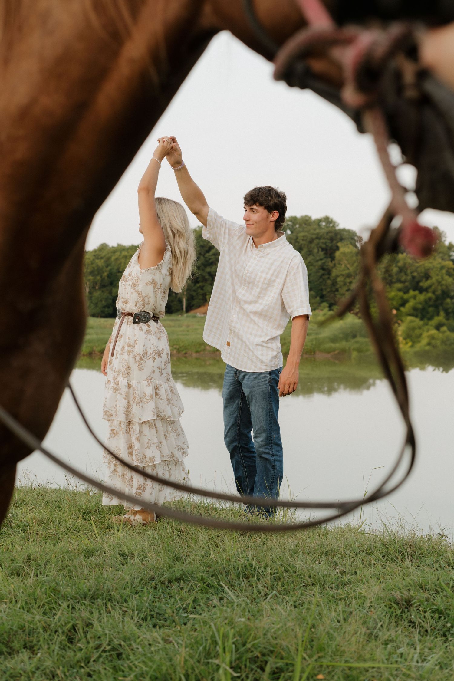A couple dances near a pond framed by horses, woman in floral dress, man in jeans and white shirt.