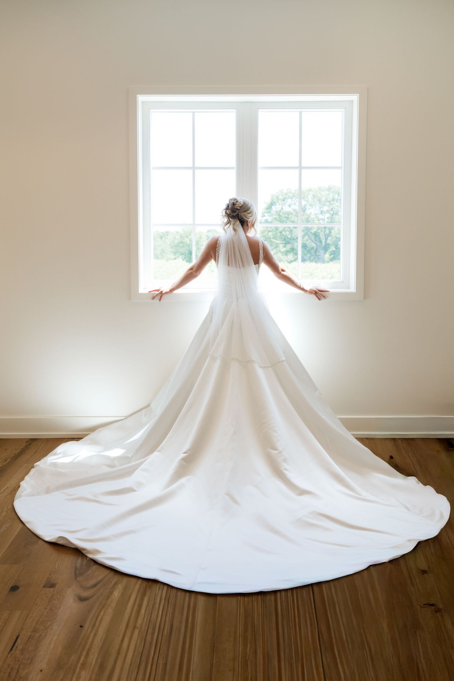 Bride in elegant white wedding gown with long train stands facing window, veil and floral hair accessory visible.