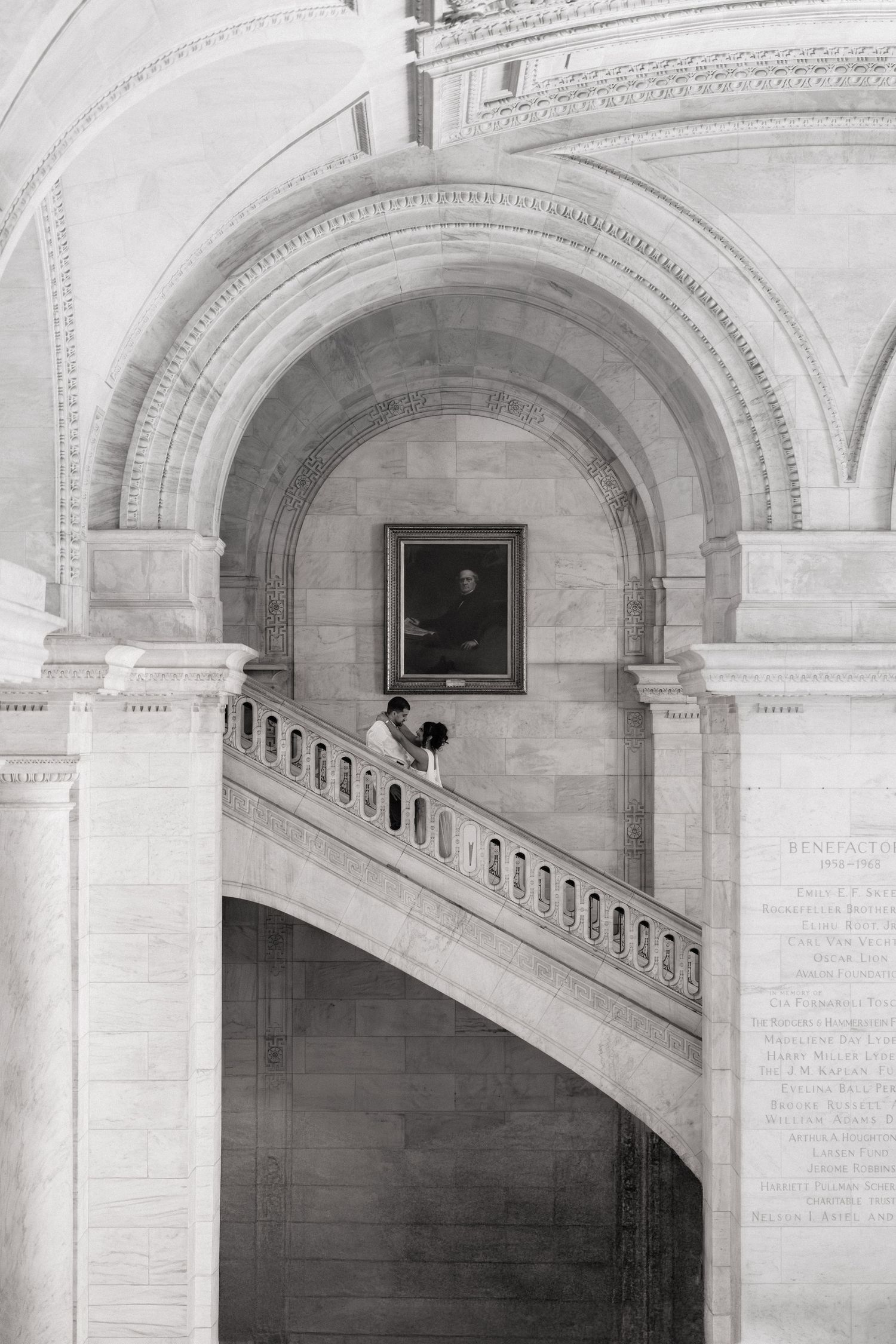 Black and white photo of a couple on grand staircase inside ornate neoclassical building with arched ceilings.