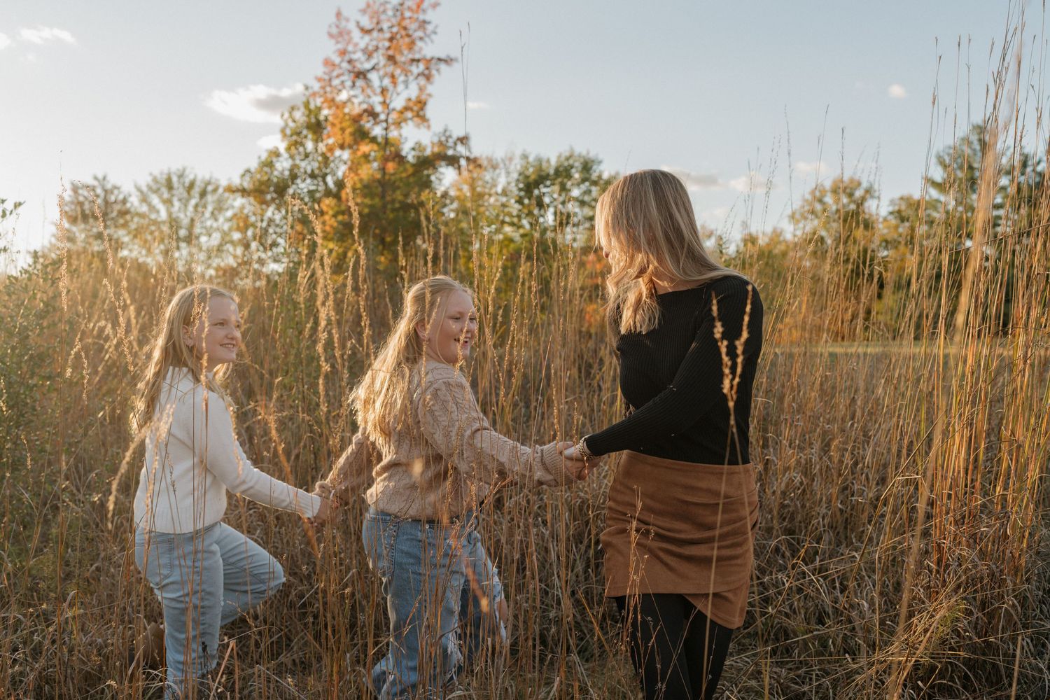 Three people hold hands and dance in a golden field at sunset with tall trees in the background.