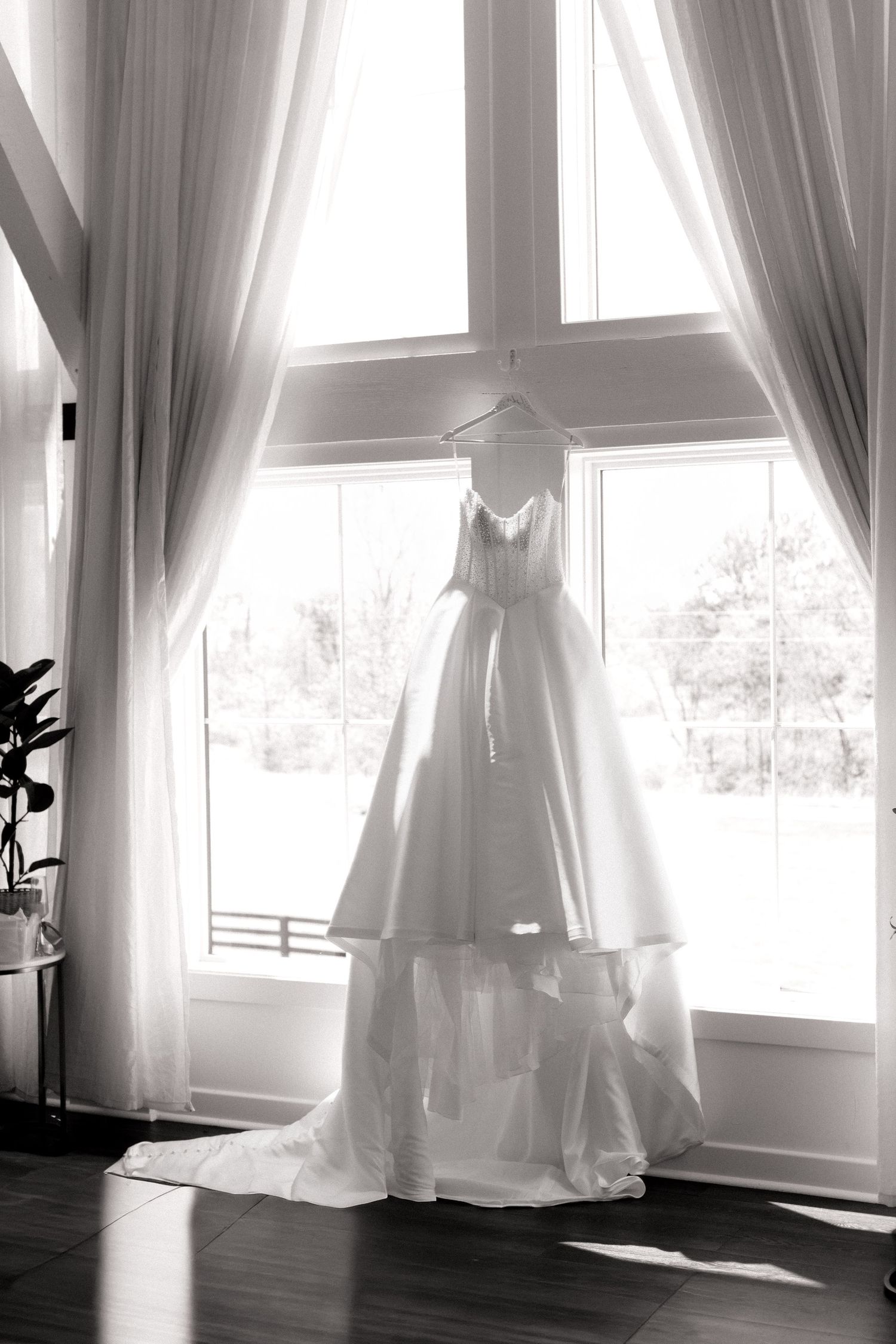 Elegant white wedding gown hanging on a hanger in front of a large window, black and white photography.
