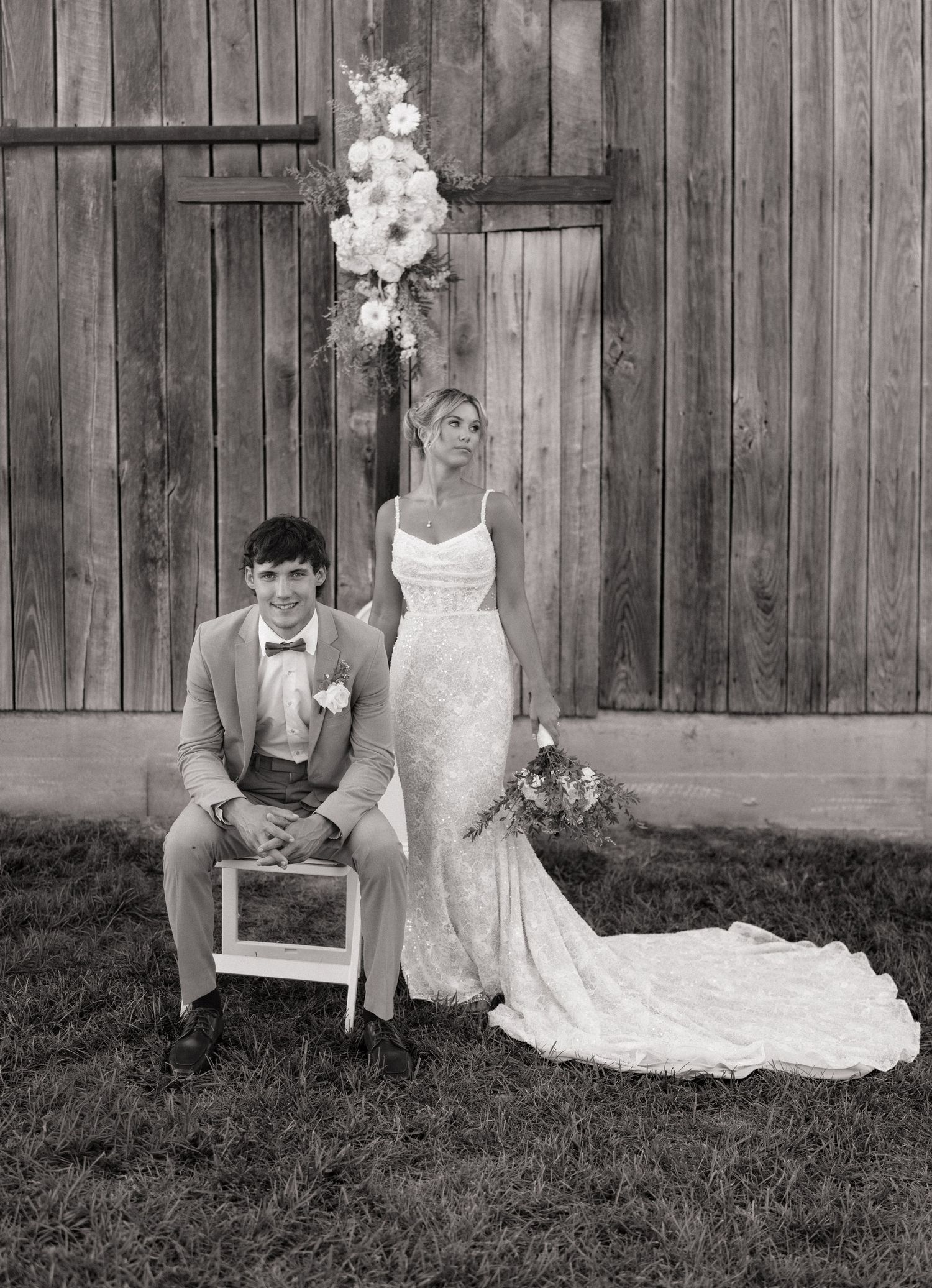 Black and white photo of bride in lace gown and groom in suit posing before rustic barn doors with floral arrangement.