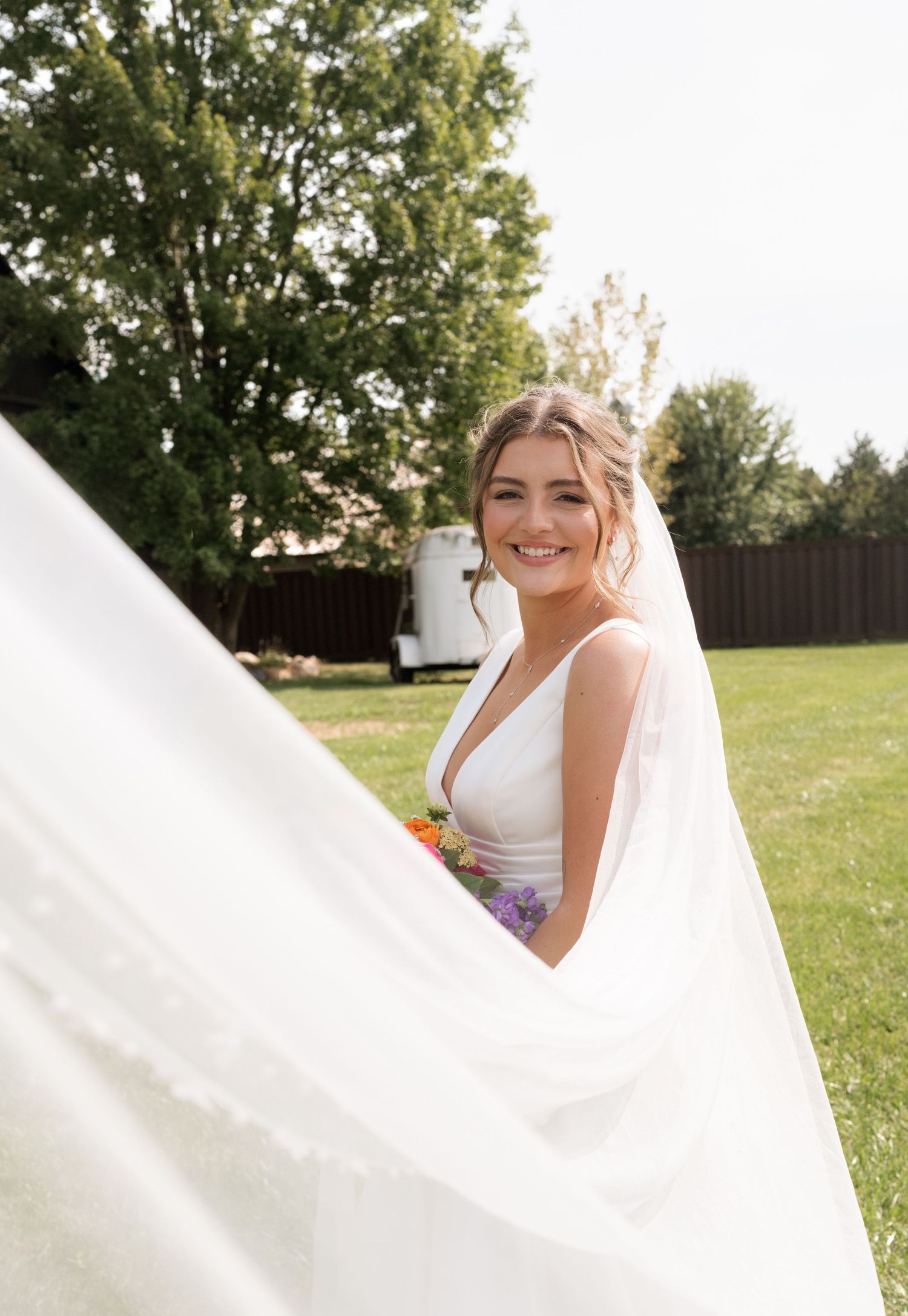 Smiling bride in a white gown holding colorful flowers, with her veil flowing in the breeze outdoors.