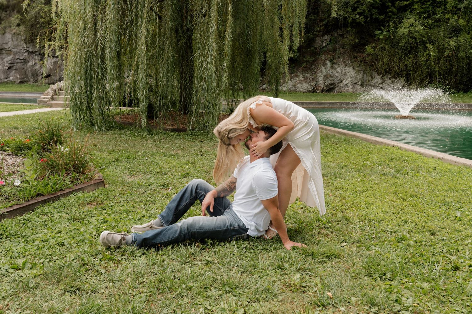 Playful couple embracing on grass near fountain and willow tree.
