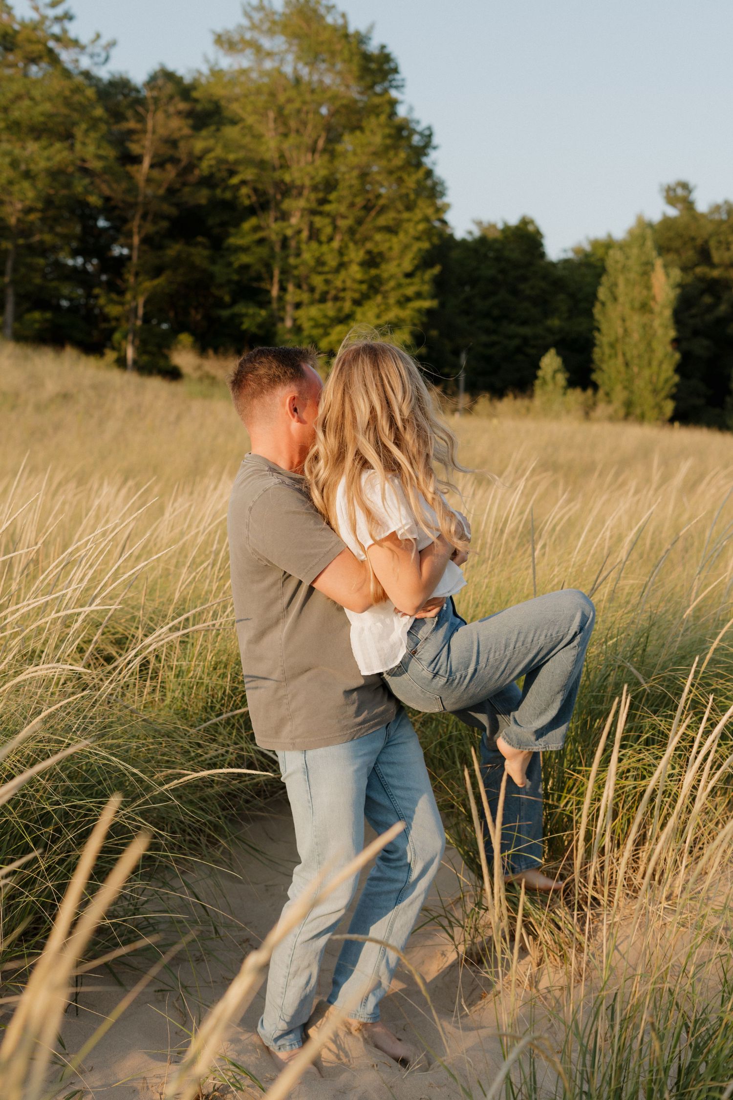 Playful couple dancing and embracing on sandy coastal path with beach grass at sunset.