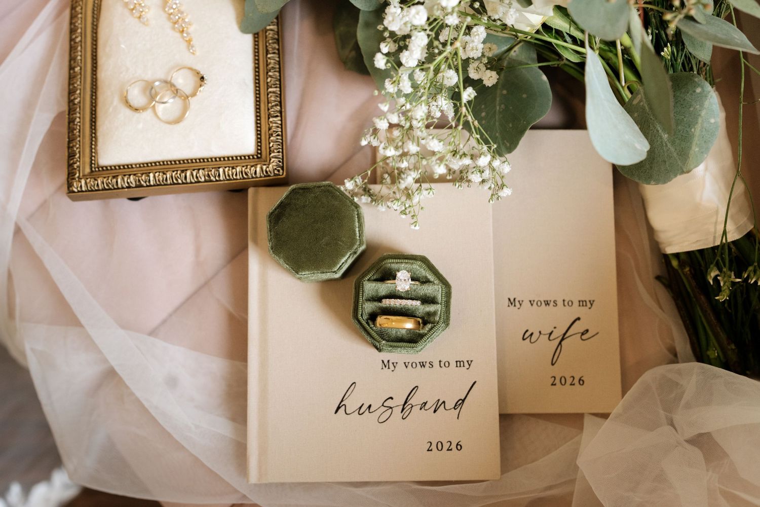 Wedding vow books for husband and wife displayed with gold rings in velvet box, eucalyptus greenery, and delicate baby's breath flowers.