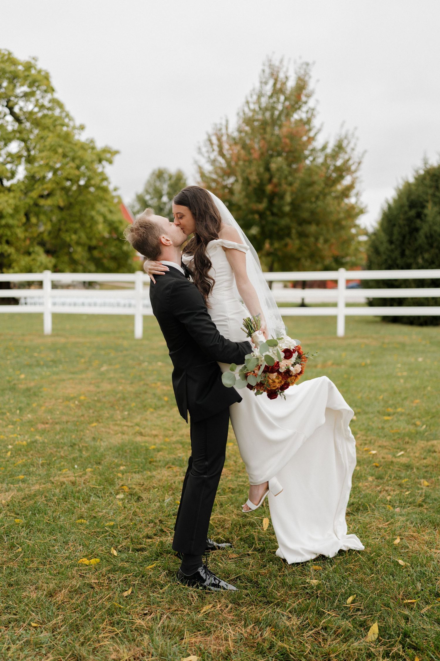 Romantic wedding couple dips and kisses on a grassy field with white fence and autumn trees in background.