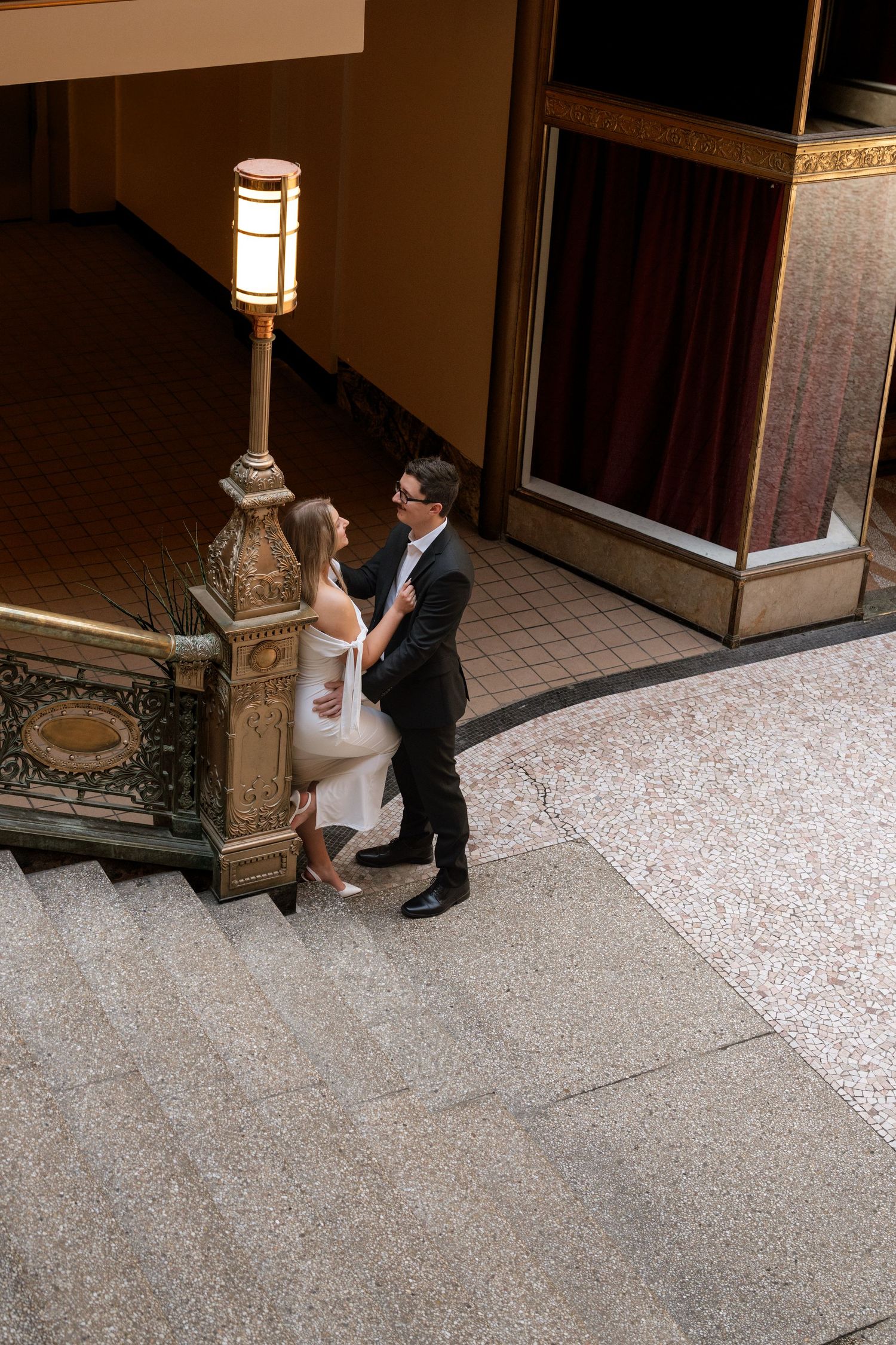 Couple in formal attire sharing a romantic moment on grand staircase near ornate lamp post in elegant venue.