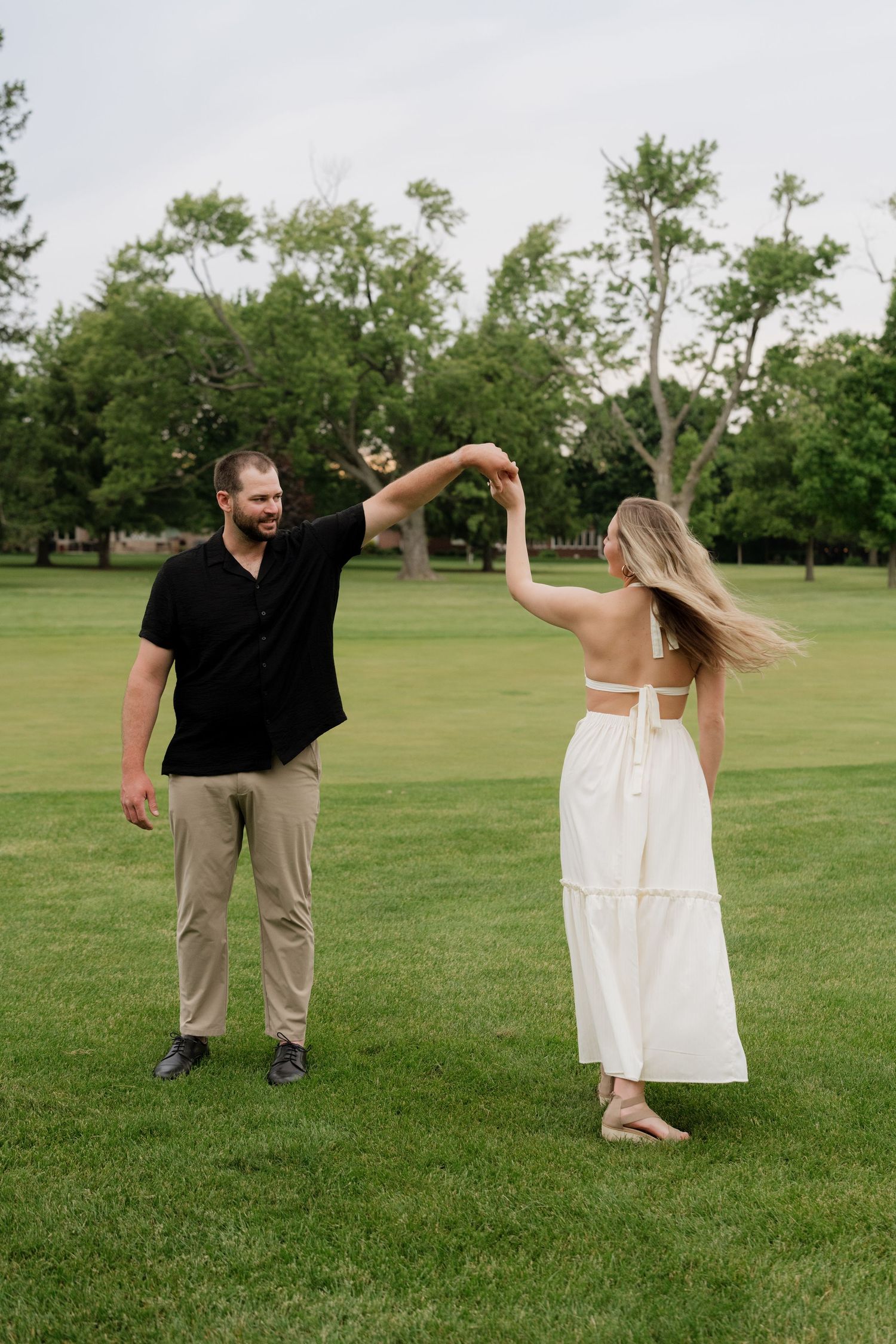 A couple dances together on a green lawn, man in black shirt and woman in white dress, with trees in background.
