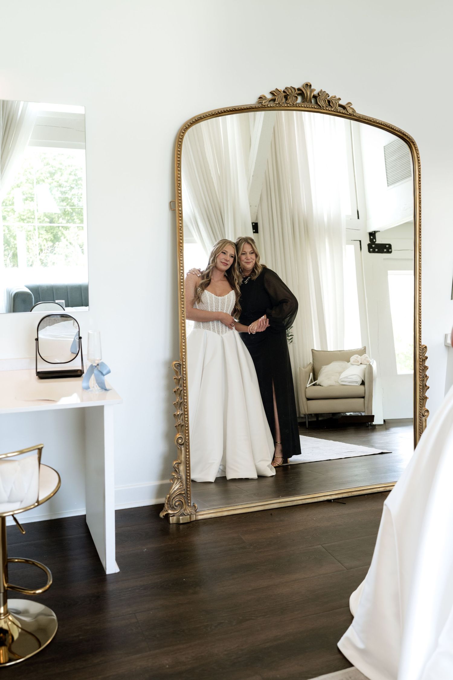 Bride and her mother share a tender moment reflected in an ornate gold floor mirror in a bright bridal suite.