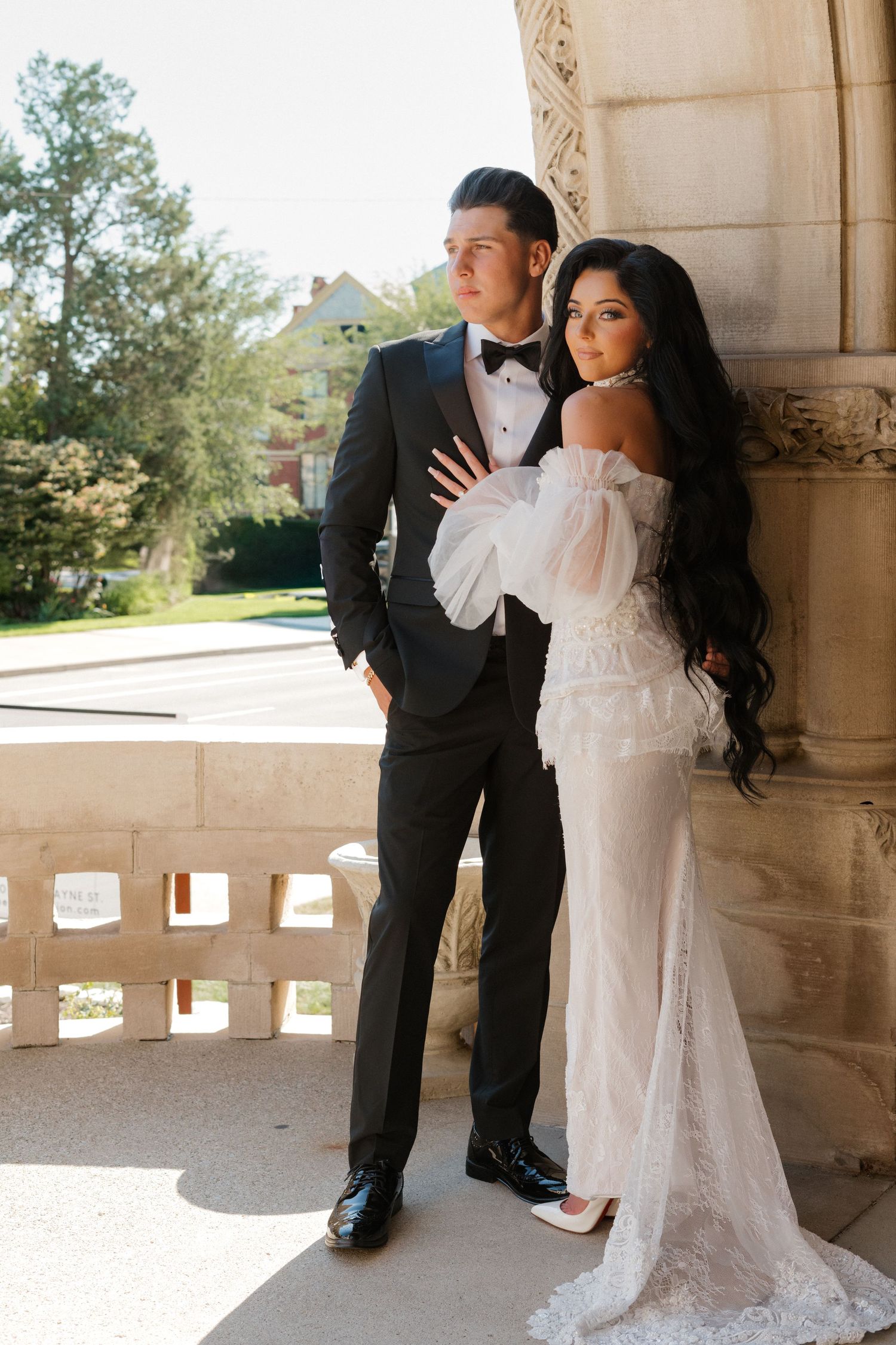 Bride in lace gown and groom in tuxedo pose on historic veranda.