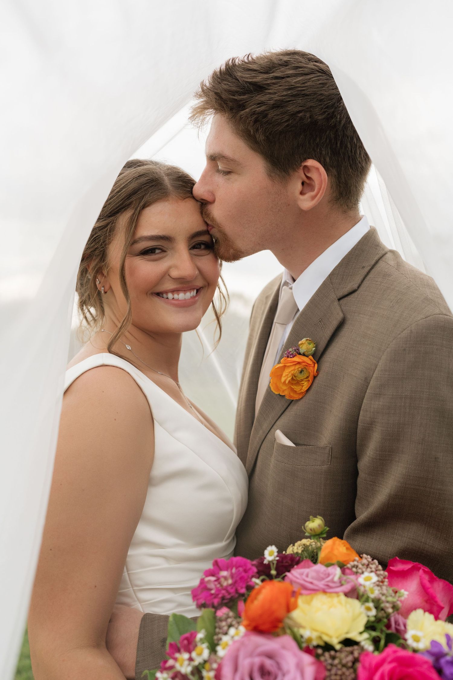Bride smiling as groom kisses her cheek under veil, holding colorful bouquet of pink, orange, and yellow roses.