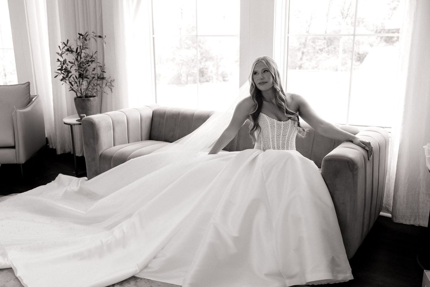 Black and white photo of a bride in a ballgown wedding dress seated gracefully on a sofa near bright windows.