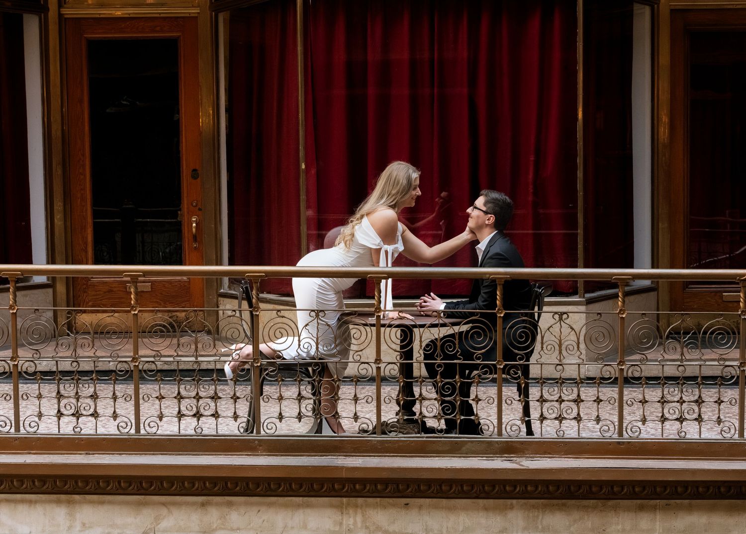 Couple sharing a romantic moment on an elegant balcony with ornate railings and a red curtain backdrop.
