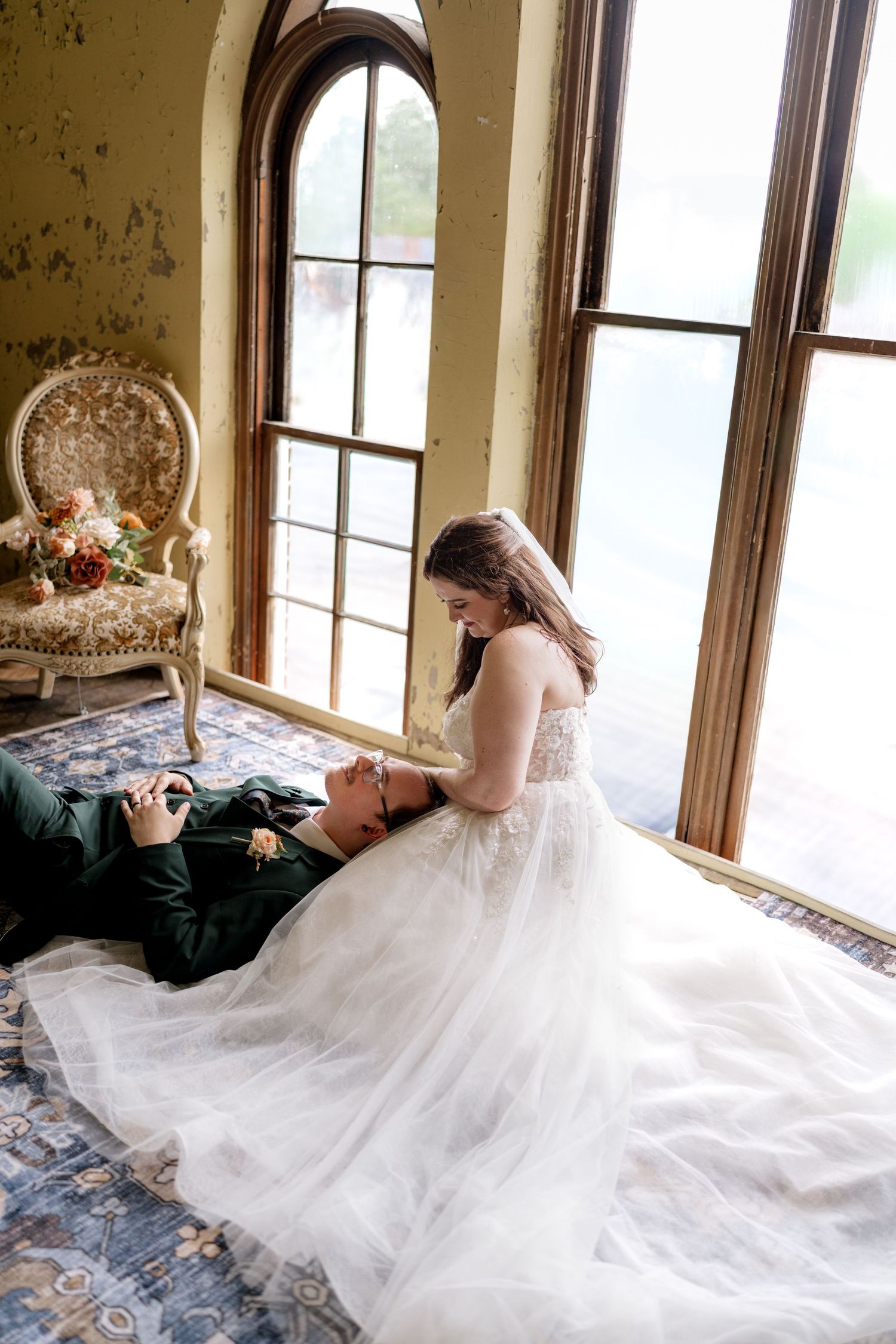 Bride in white tulle gown sits on floor with groom in green suit lying in her lap near large arched windows.