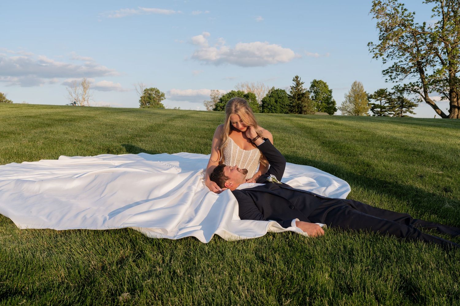 Wedding couple relaxing on white bridal gown spread across lush green meadow at golden hour sunset.