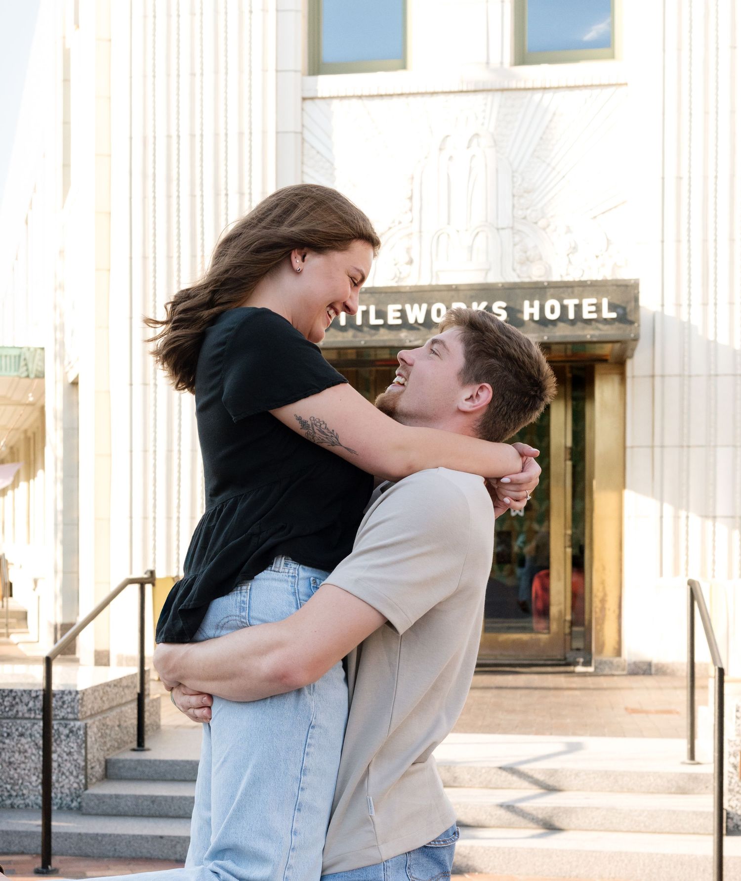 Couple embracing and laughing outside the Bottleworks Hotel, man lifting woman in a joyful romantic moment.