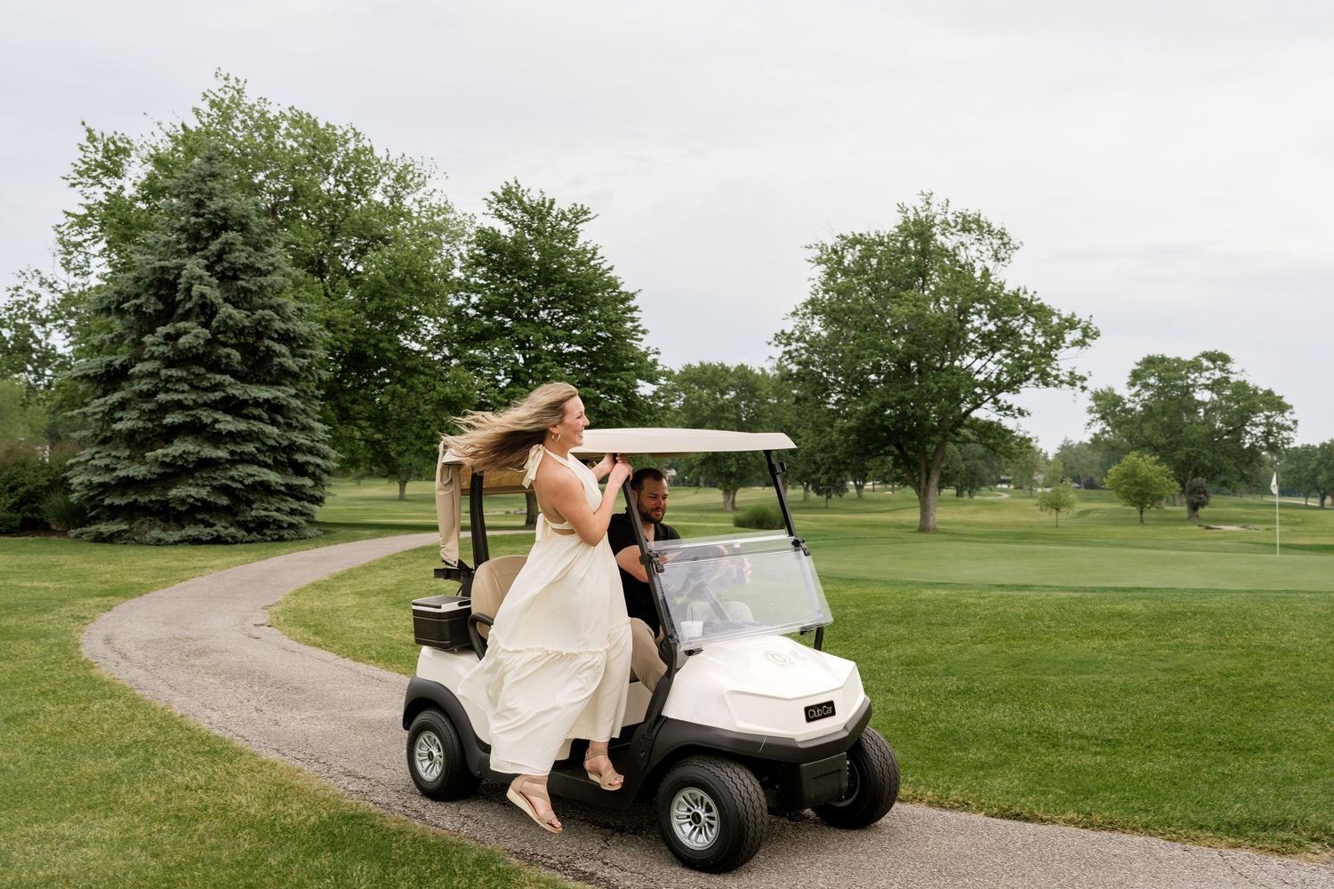 Bride and groom riding a white golf cart on a scenic golf course path surrounded by lush green trees.