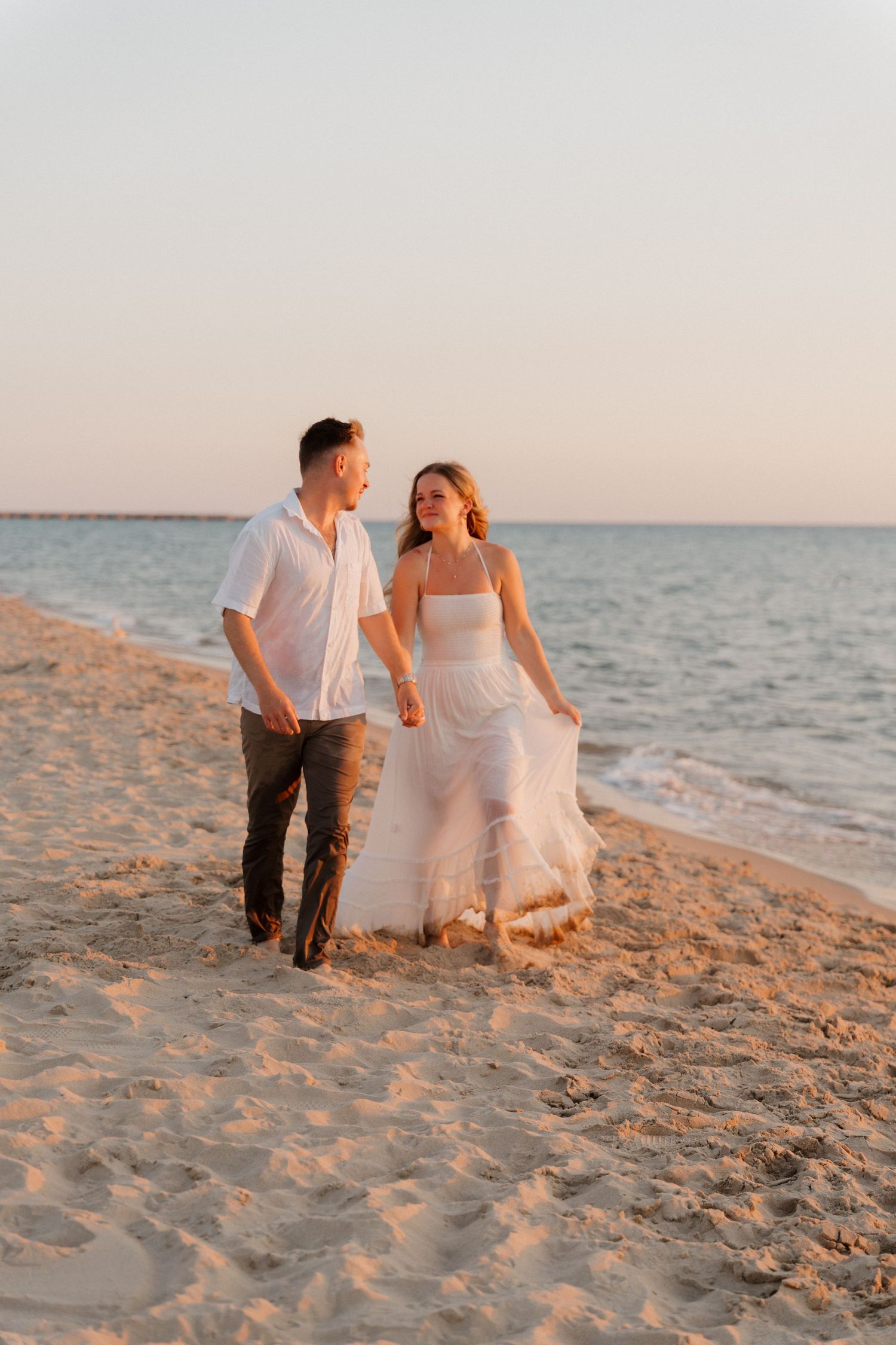 Couple walks hand in hand on sandy beach at golden sunset, woman in flowing white dress, man in white shirt.