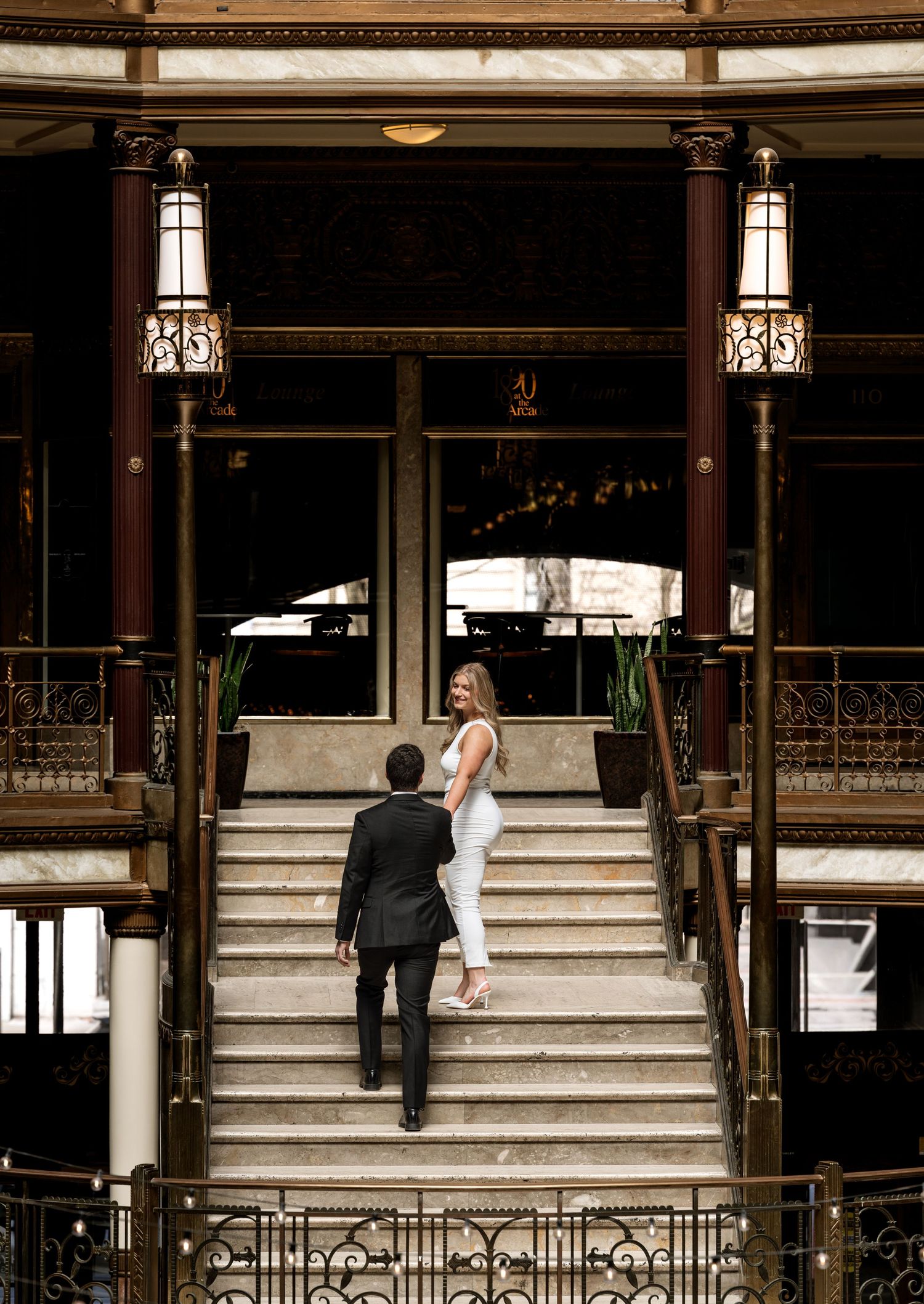 Couple walking up grand marble staircase in elegant historic building, woman in white outfit glancing back at camera.