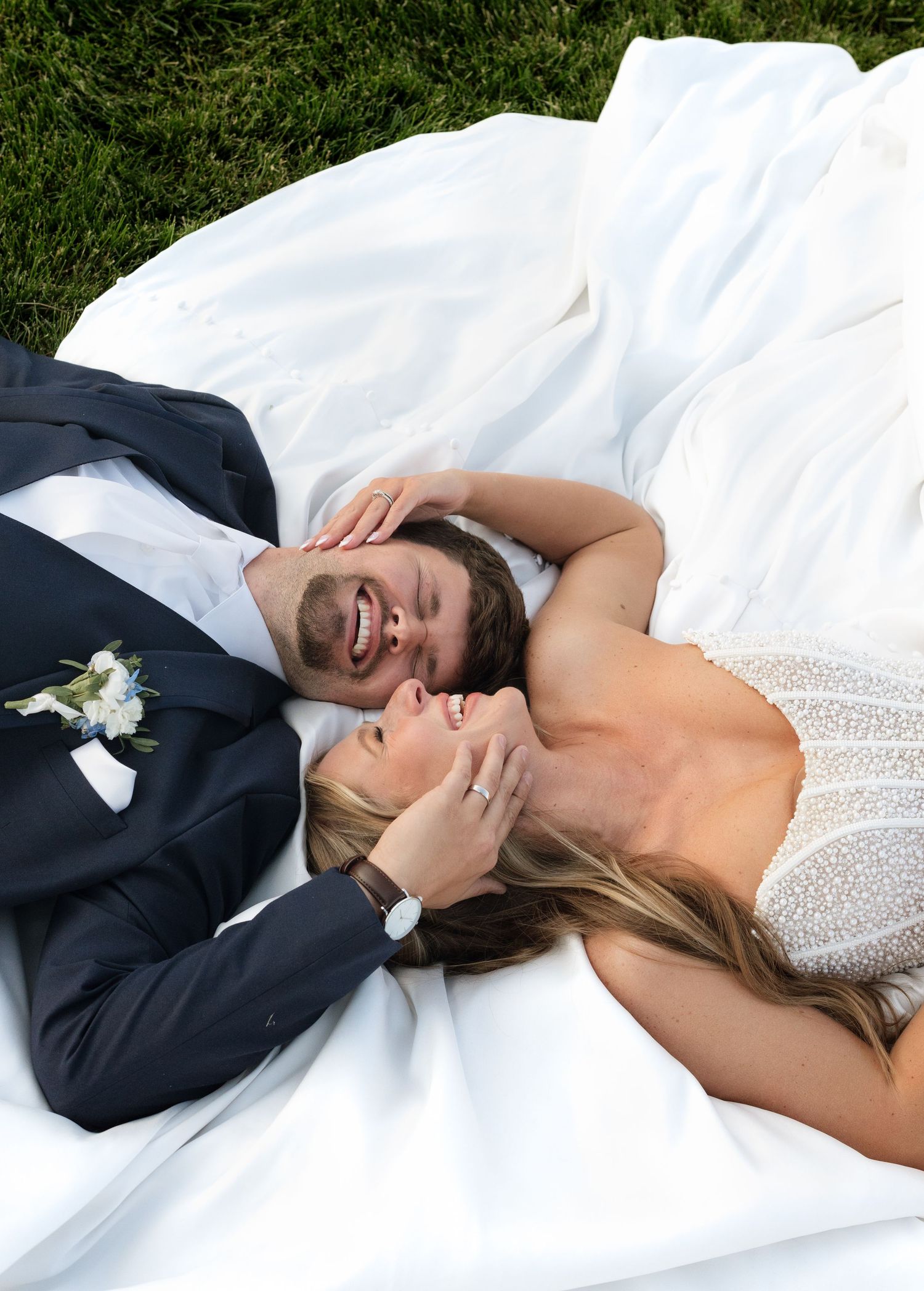 Bride and groom lying on white wedding dress in green grass, laughing and sharing a joyful moment together.