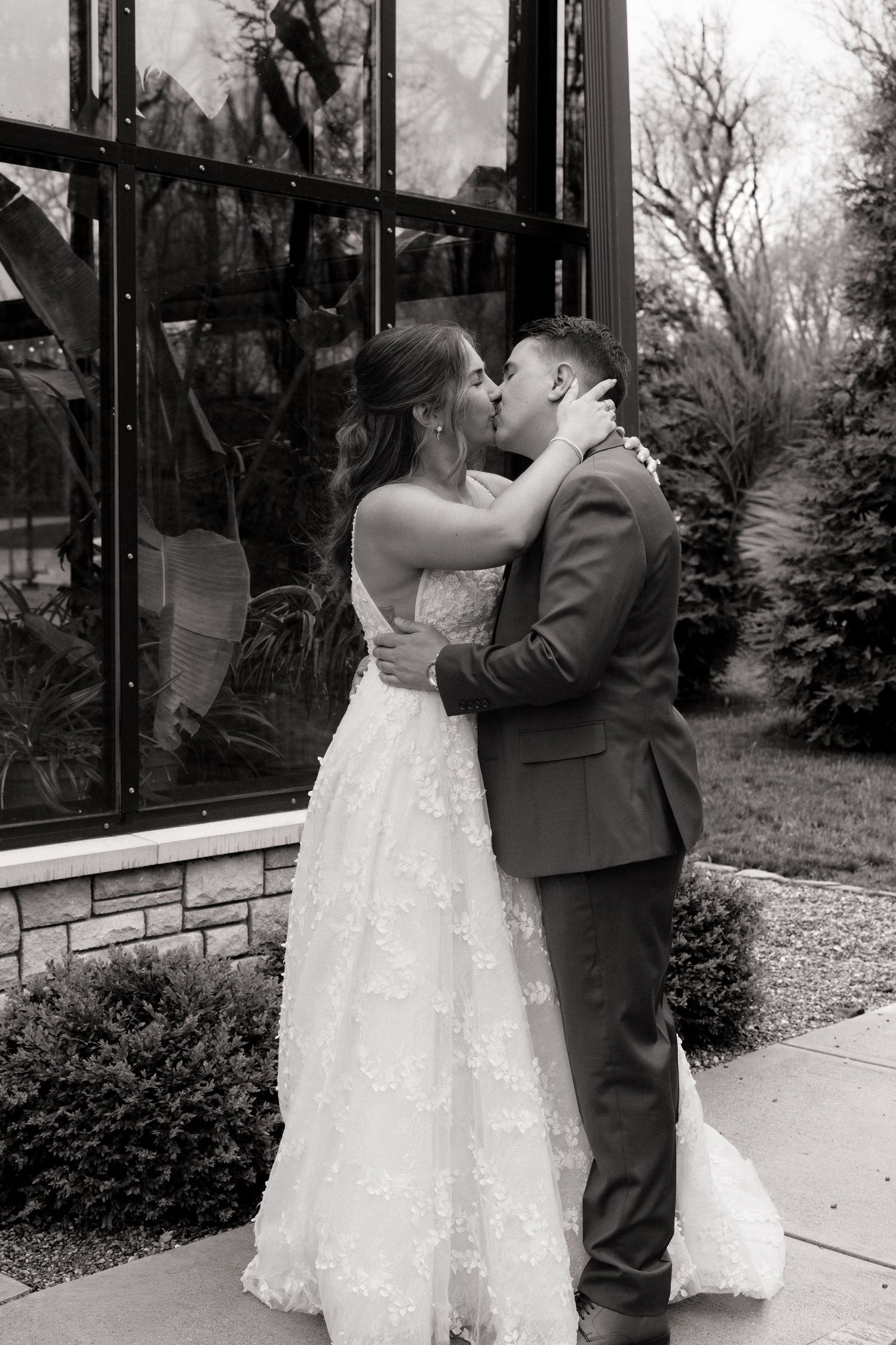 Black and white photo of a bride and groom sharing a kiss outside a glass greenhouse on their wedding day.