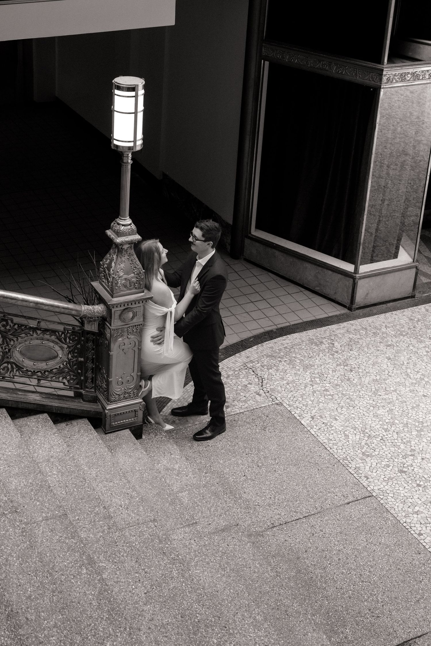 Black and white aerial view of a couple holding hands near an ornate street lamp outside a building.