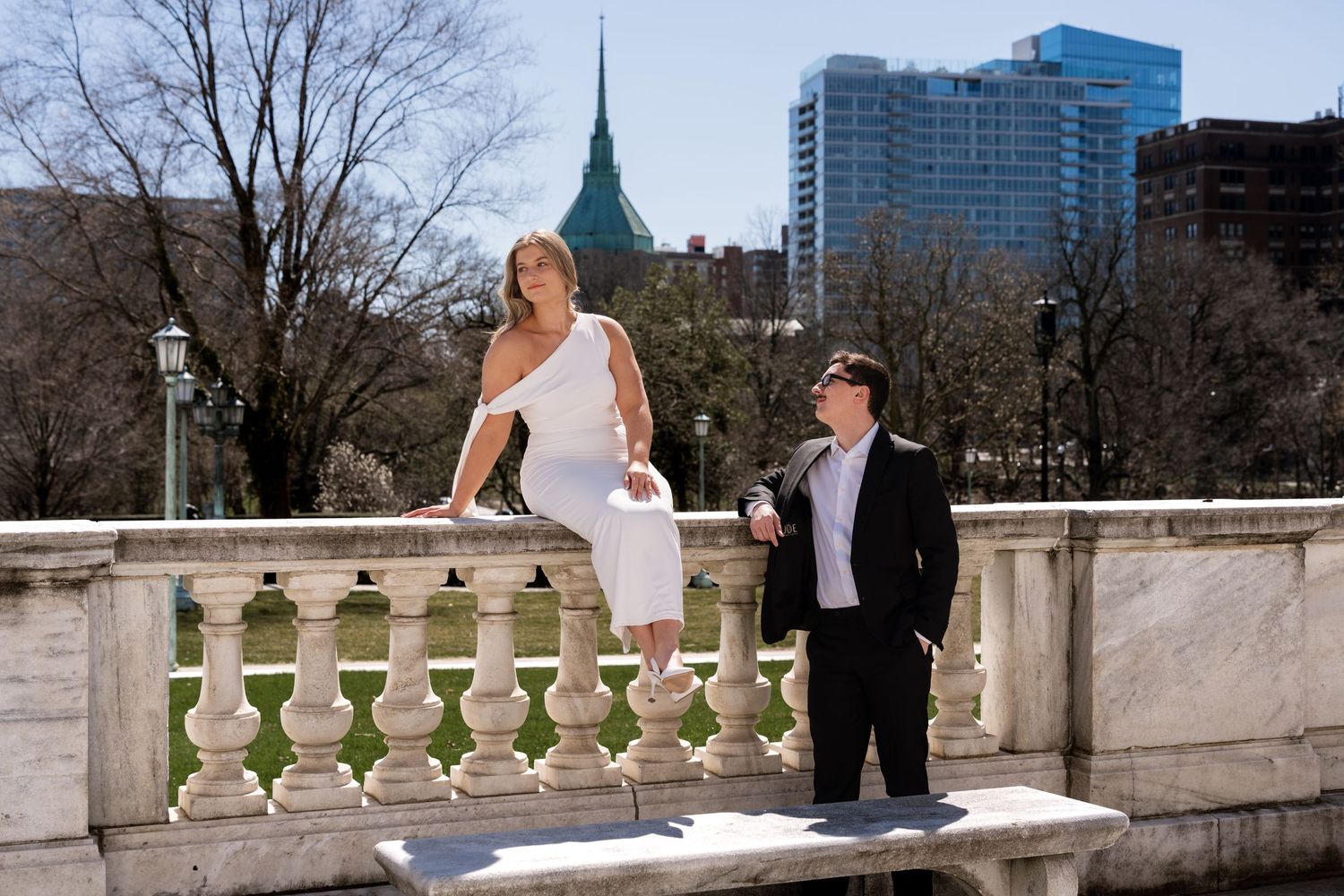 Couple posing on ornate stone balustrade with city skyline backdrop on a sunny day.