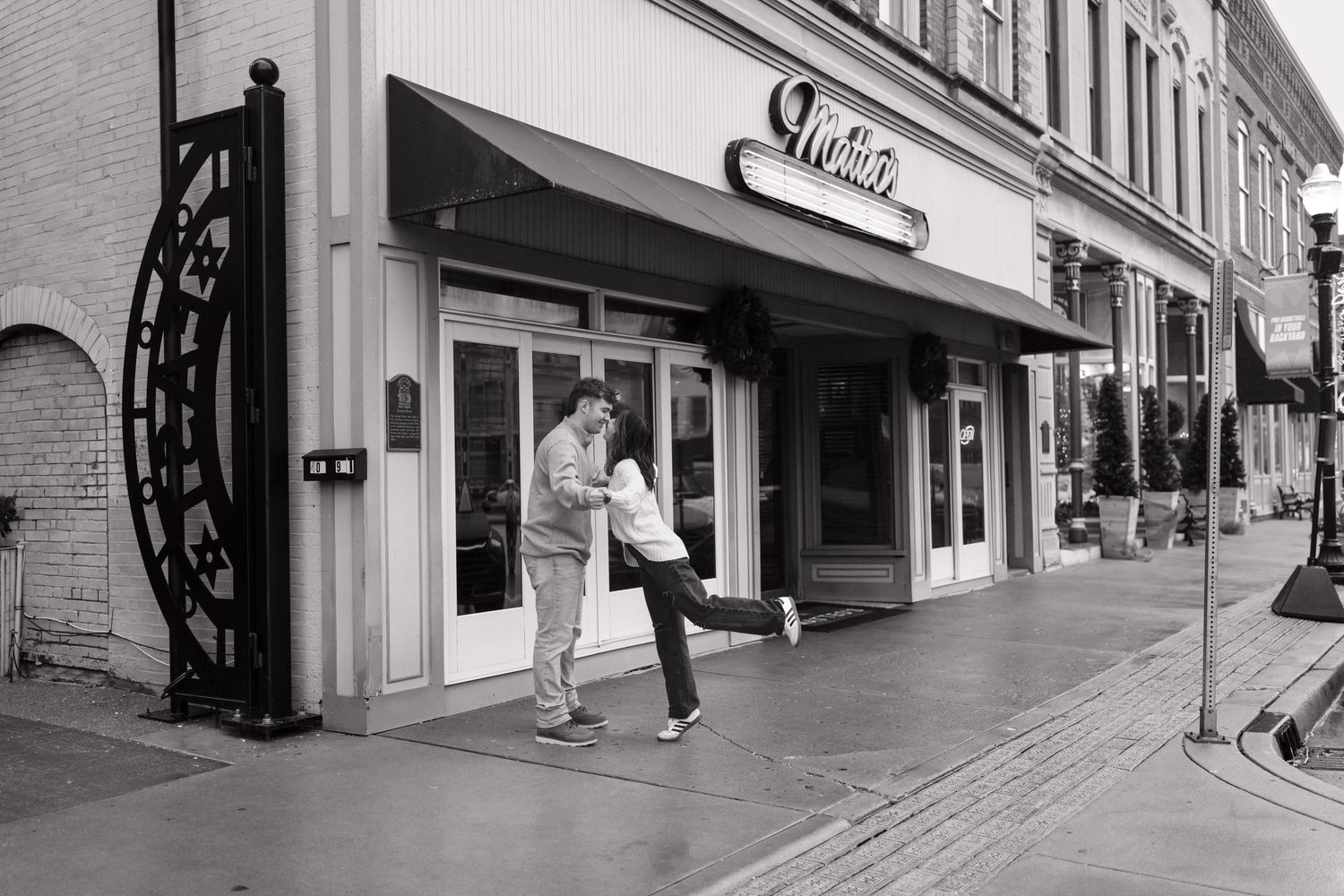 A black and white photograph shows two people dancing playfully on a sidewalk in front of a storefront.