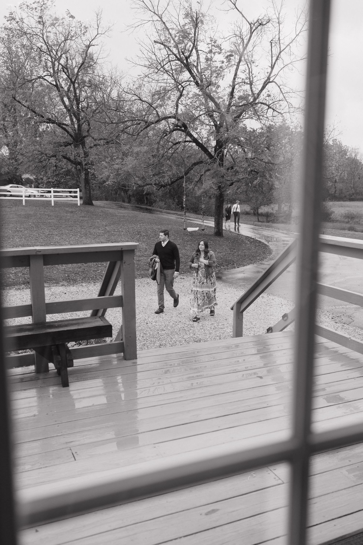 Black and white photo of couple on wooden deck through window frame with white fence in background.