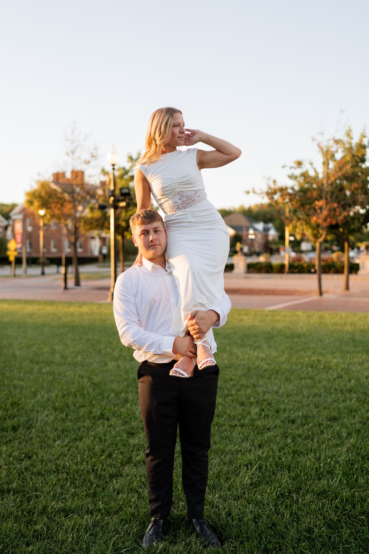 A man in white shirt lifts a woman in white dress on his shoulders in a sunny park setting.