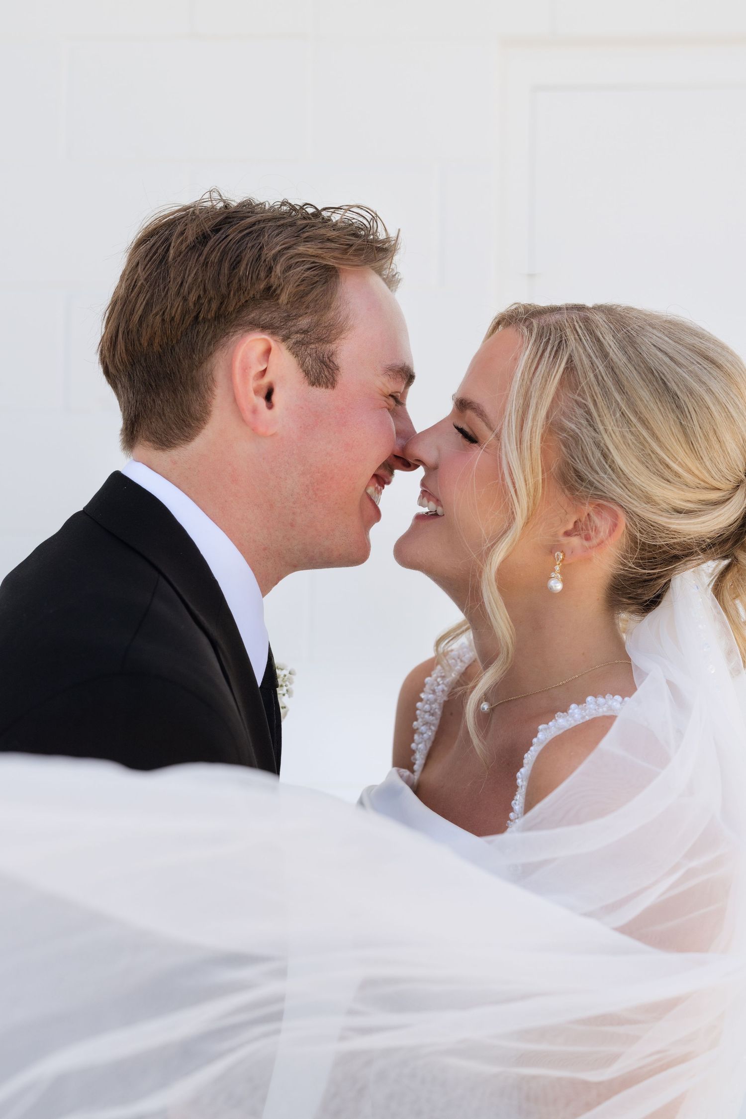 Bride and groom sharing a joyful almost-kiss moment, wrapped in a flowing white veil on their wedding day.